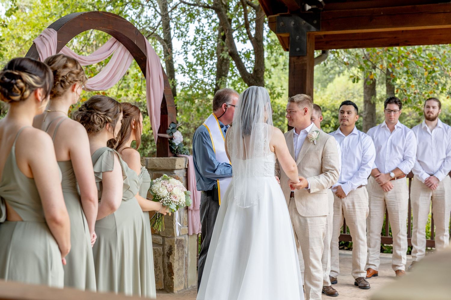 Outdoor wedding ceremony under a wooden pavilion with bridesmaids in sage green dresses and groomsmen in tan suits.