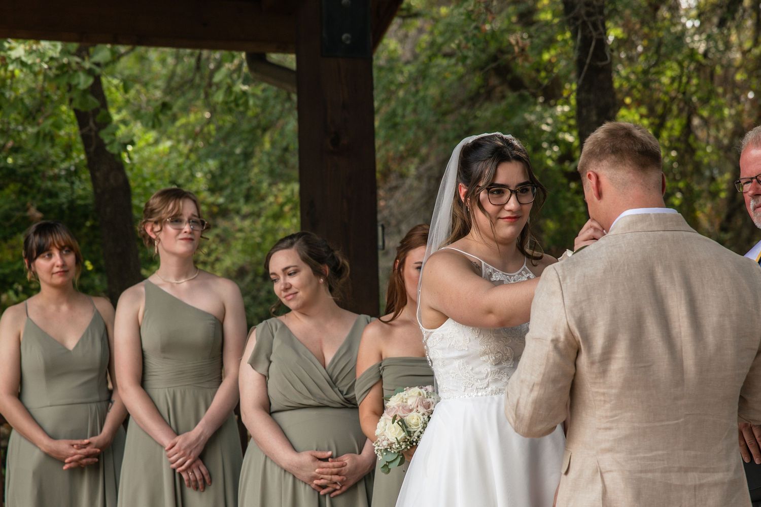 Bride in a white gown and veil faces the groom in a tan suit during an outdoor wedding ceremony as bridesmaids in sage green dresses watch.