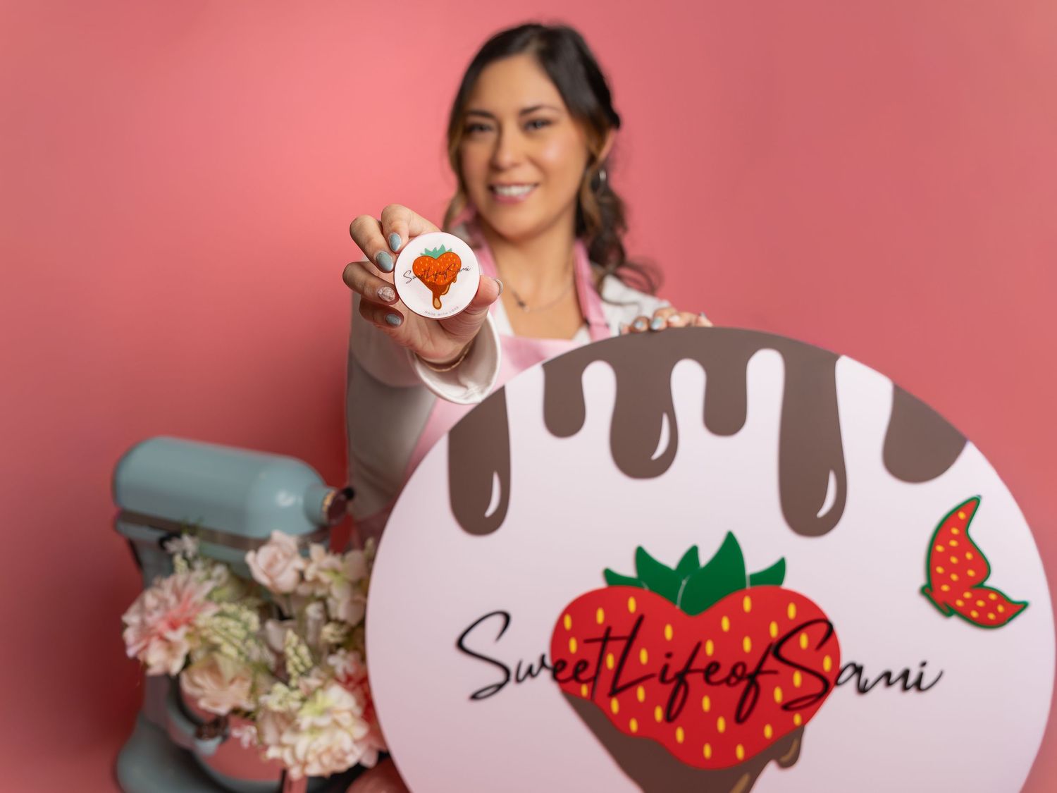 woman holding a strawberry logo button beside a Sweet Life of Sami sign with chocolate drizzle, a pink stand mixer, and flowers. Rhome Tx