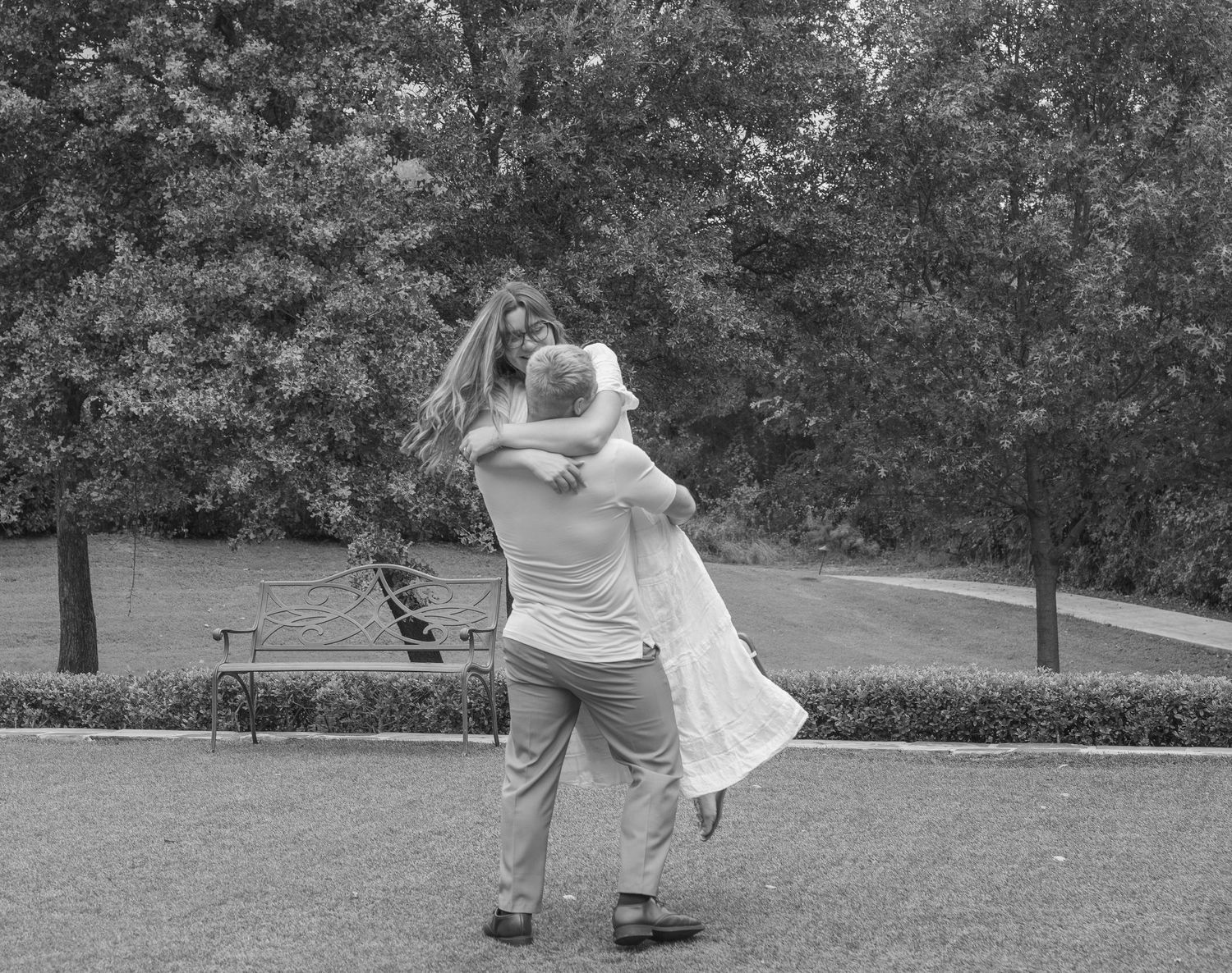 Black and white photo of a couple embracing and spinning in a park with lush trees in the background.