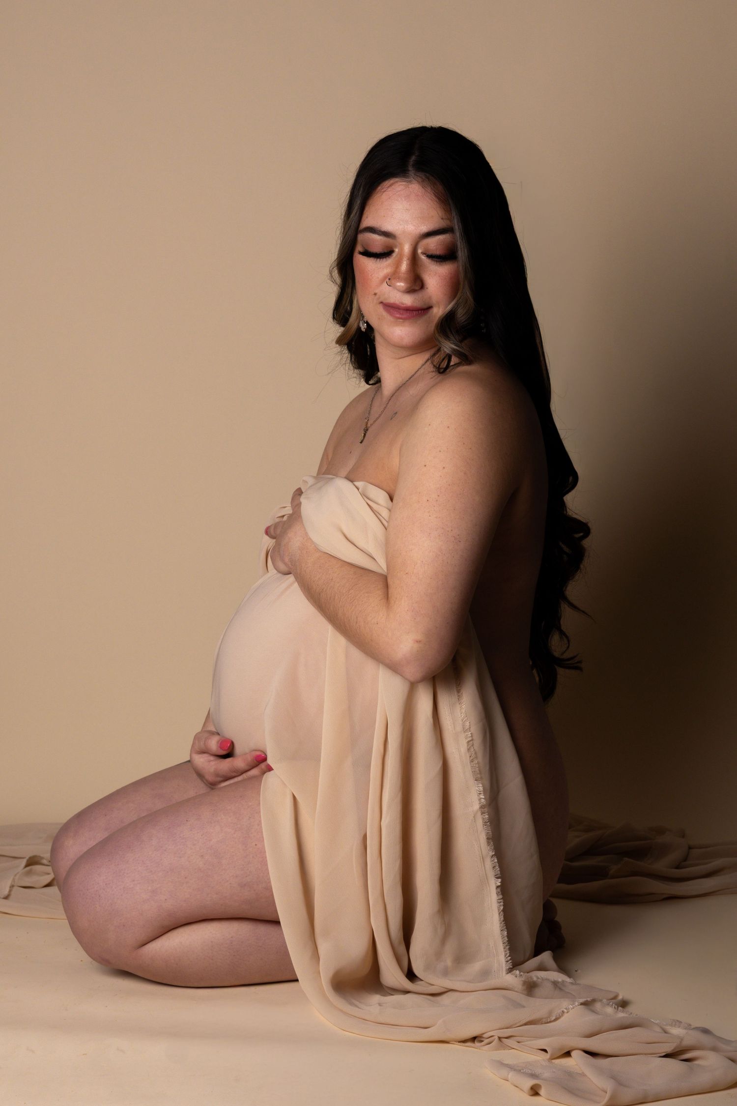 Pregnant woman kneeling in beige chiffon wrap, cradling baby bump, studio maternity portrait.