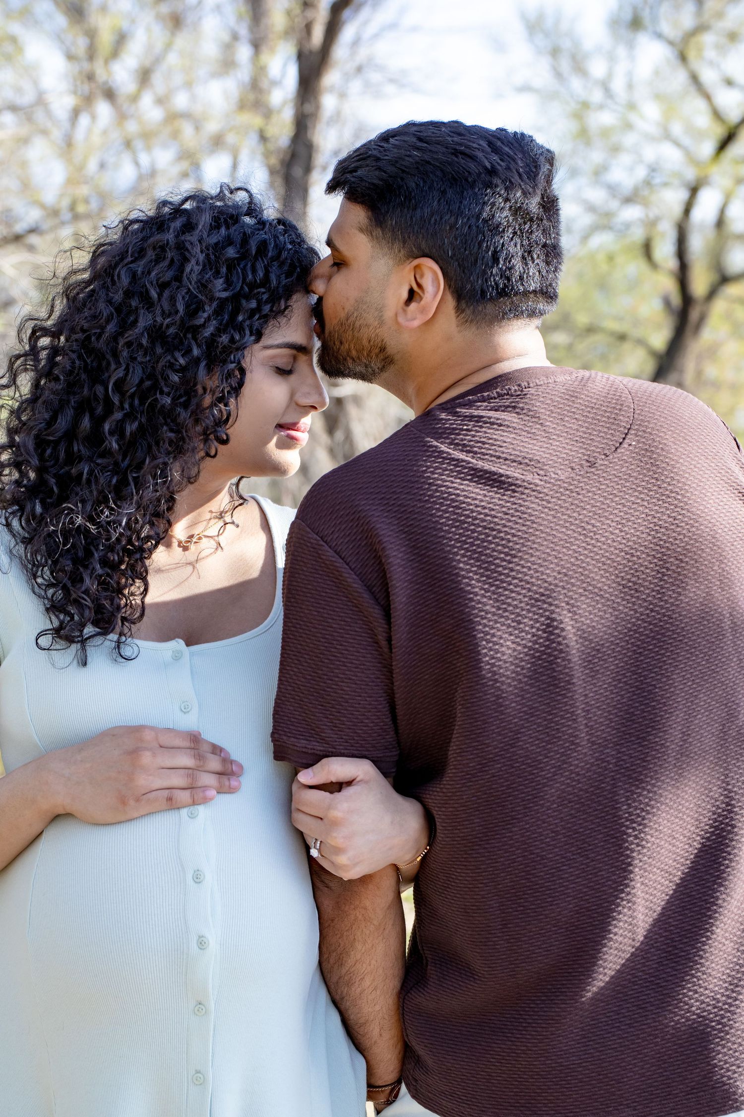 Man tenderly kissing pregnant woman on the forehead outdoors, with trees visible in the background.