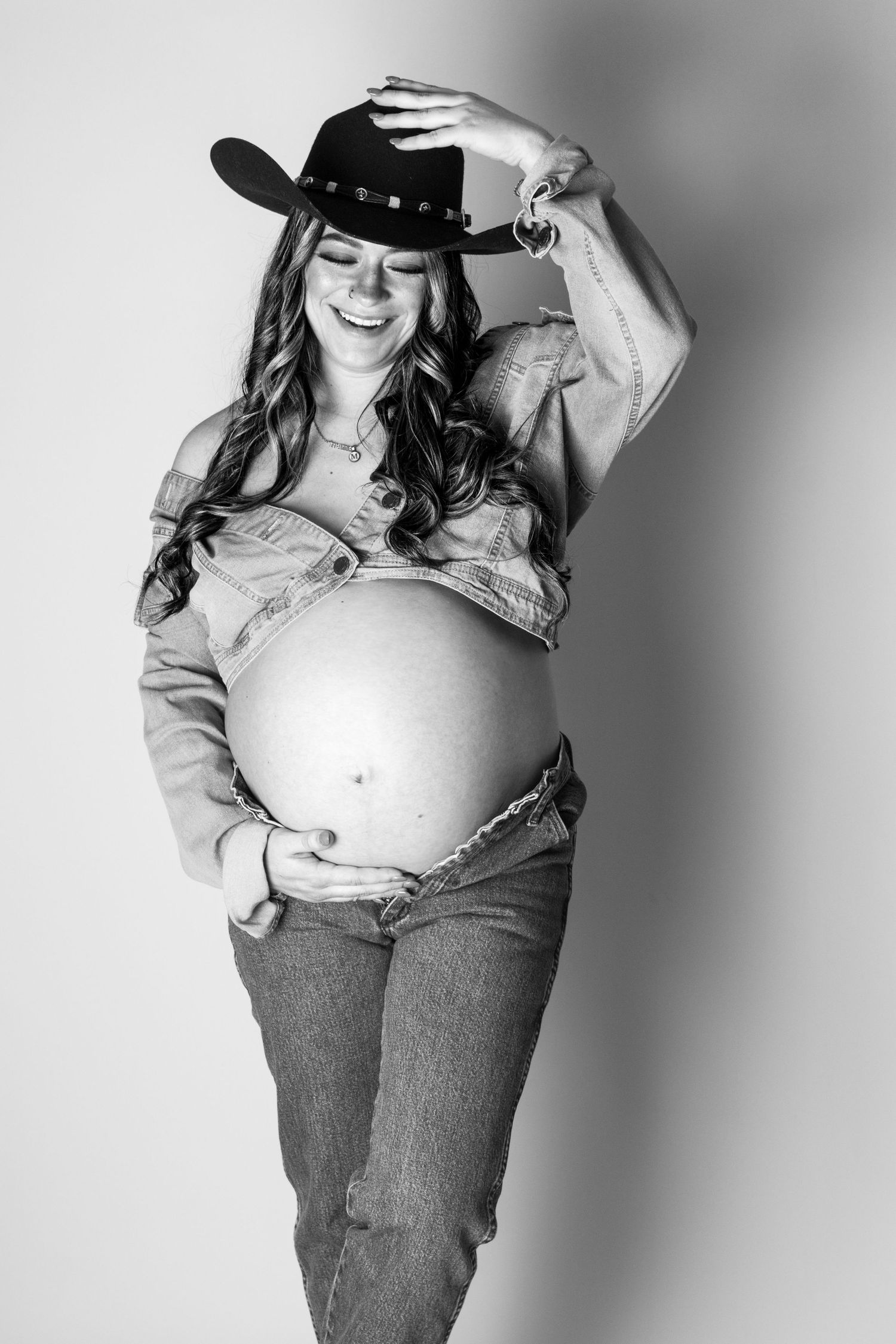 Black and white photo of a smiling pregnant woman in cowboy hat, denim jacket, and jeans cradling her baby bump.
