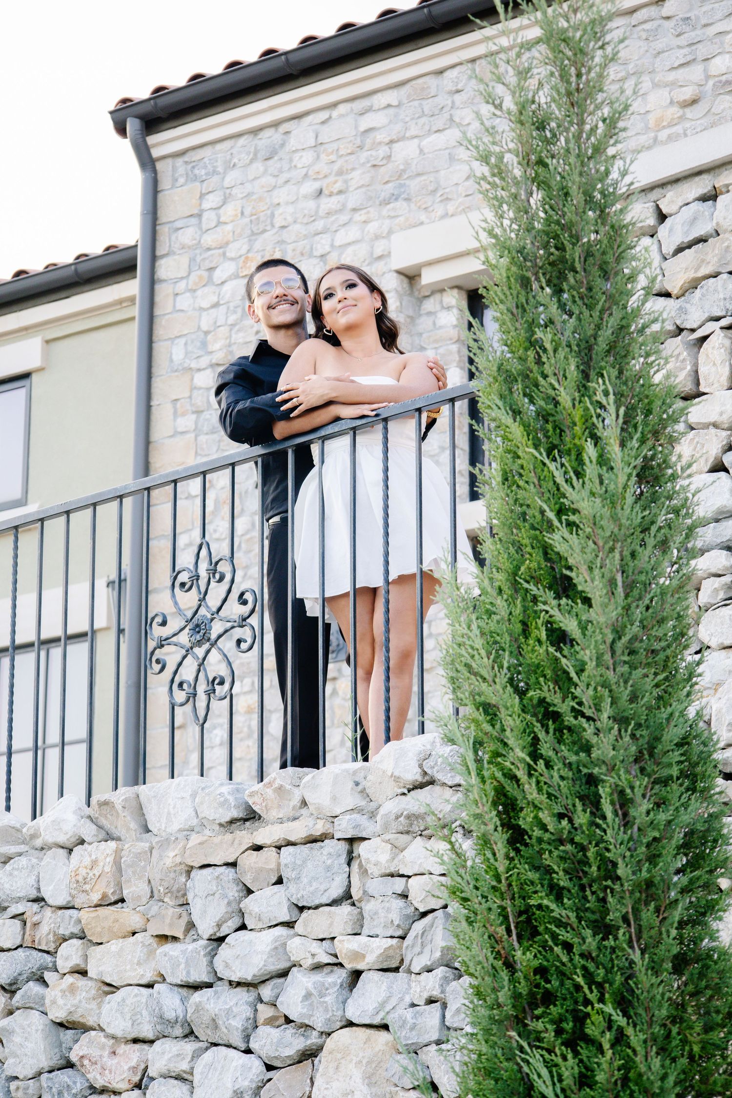 Couple embracing on a stone balcony with wrought iron railing beside a tall cypress tree at a rustic villa.