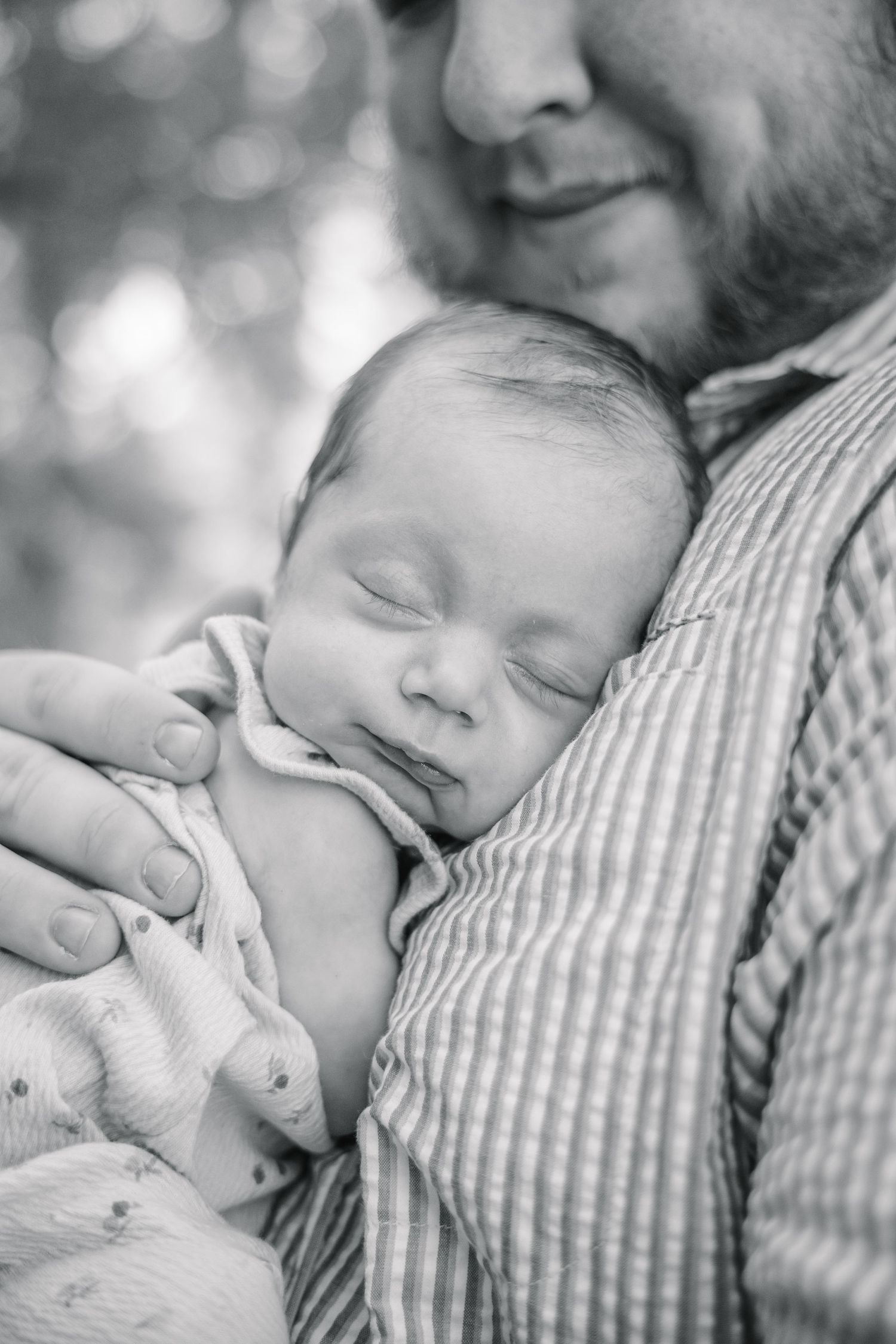 Black and white photo of a sleeping newborn baby cradled in the arms of a smiling bearded father outdoors.