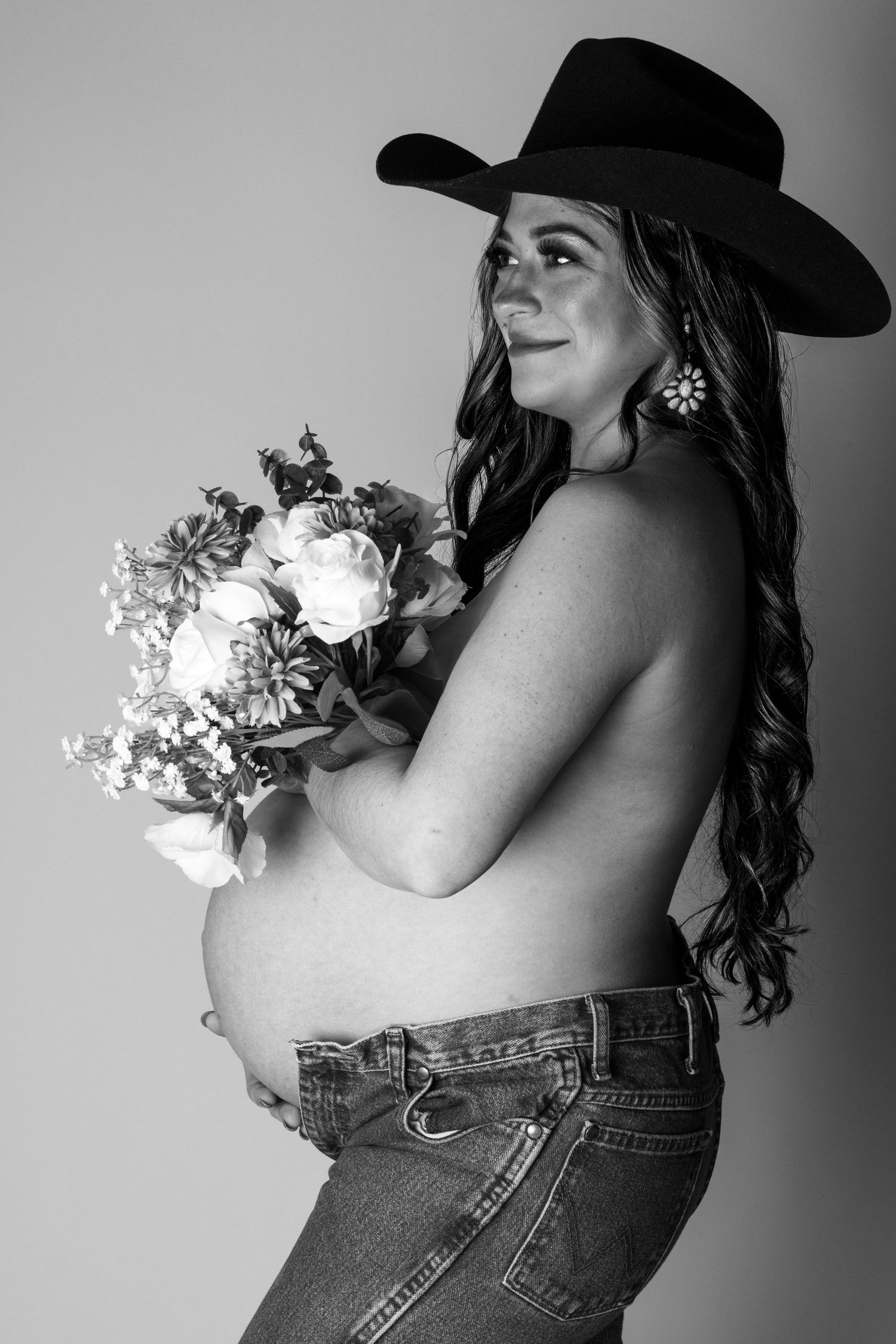 Pregnant woman in cowboy hat holding flower bouquet, showing baby bump in black and white maternity portrait.