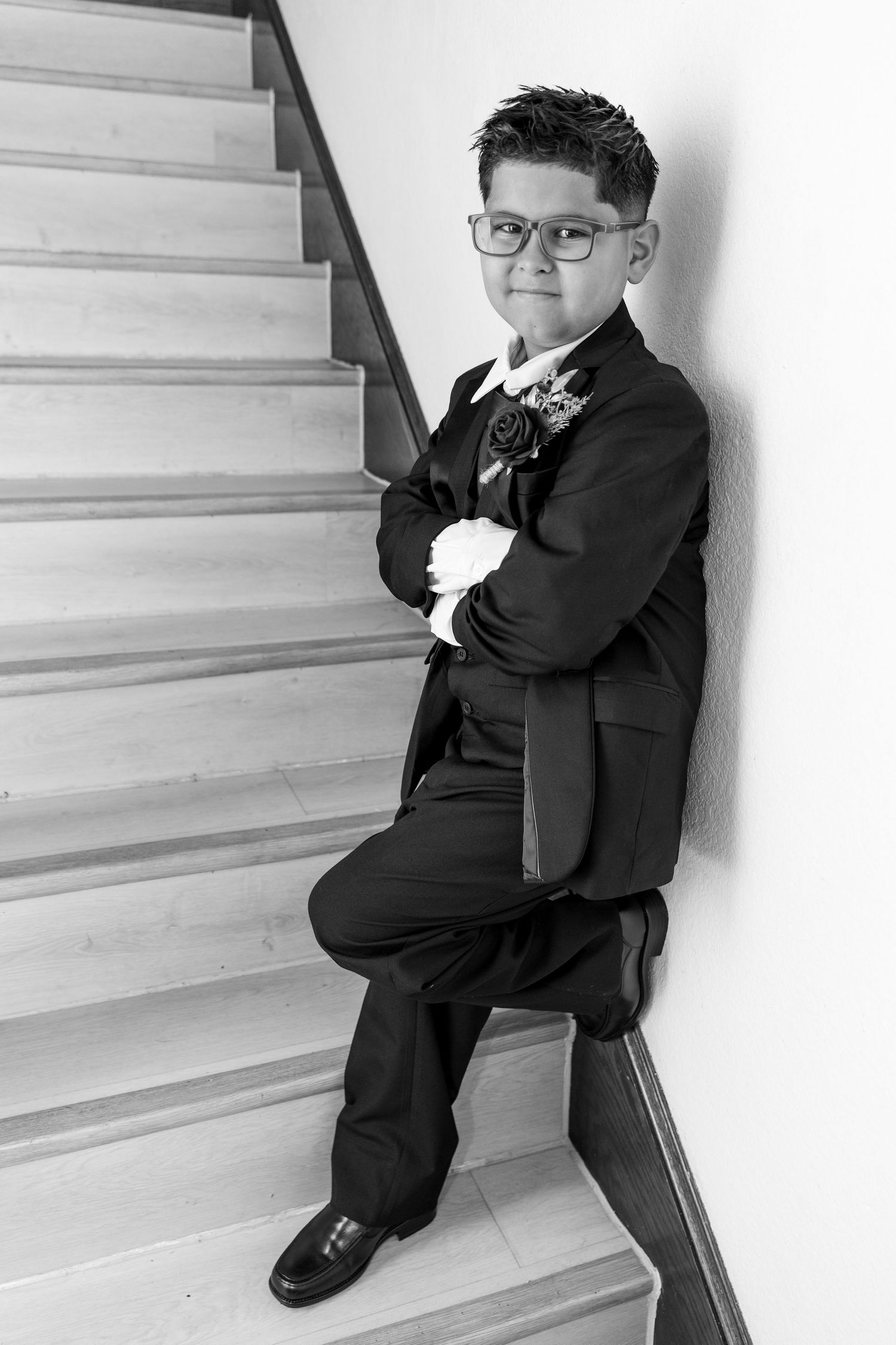 Young boy in black tuxedo with boutonniere posing confidently on staircase, black and white wedding photo.