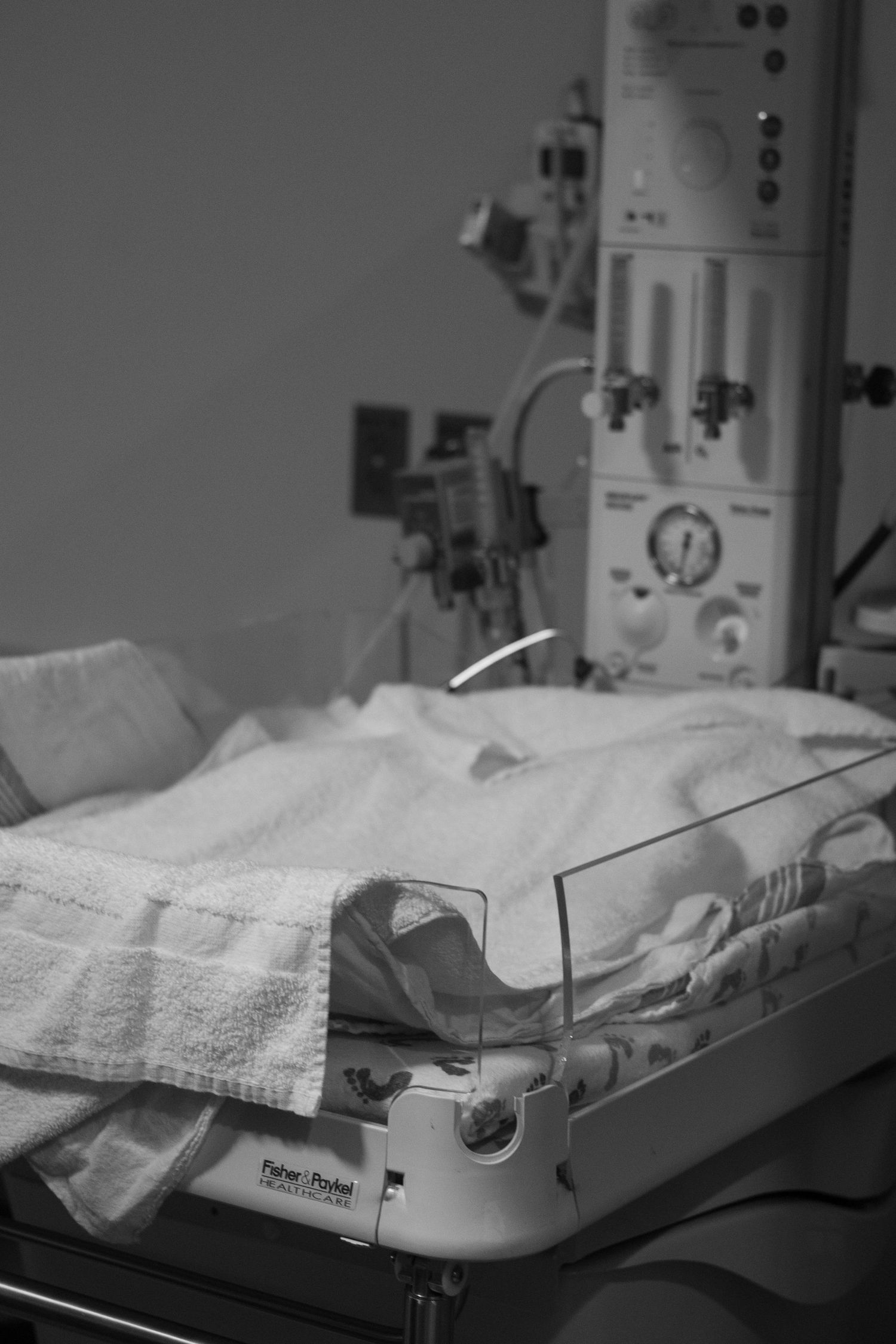 Newborn baby resting in a hospital incubator surrounded by medical equipment in black and white.