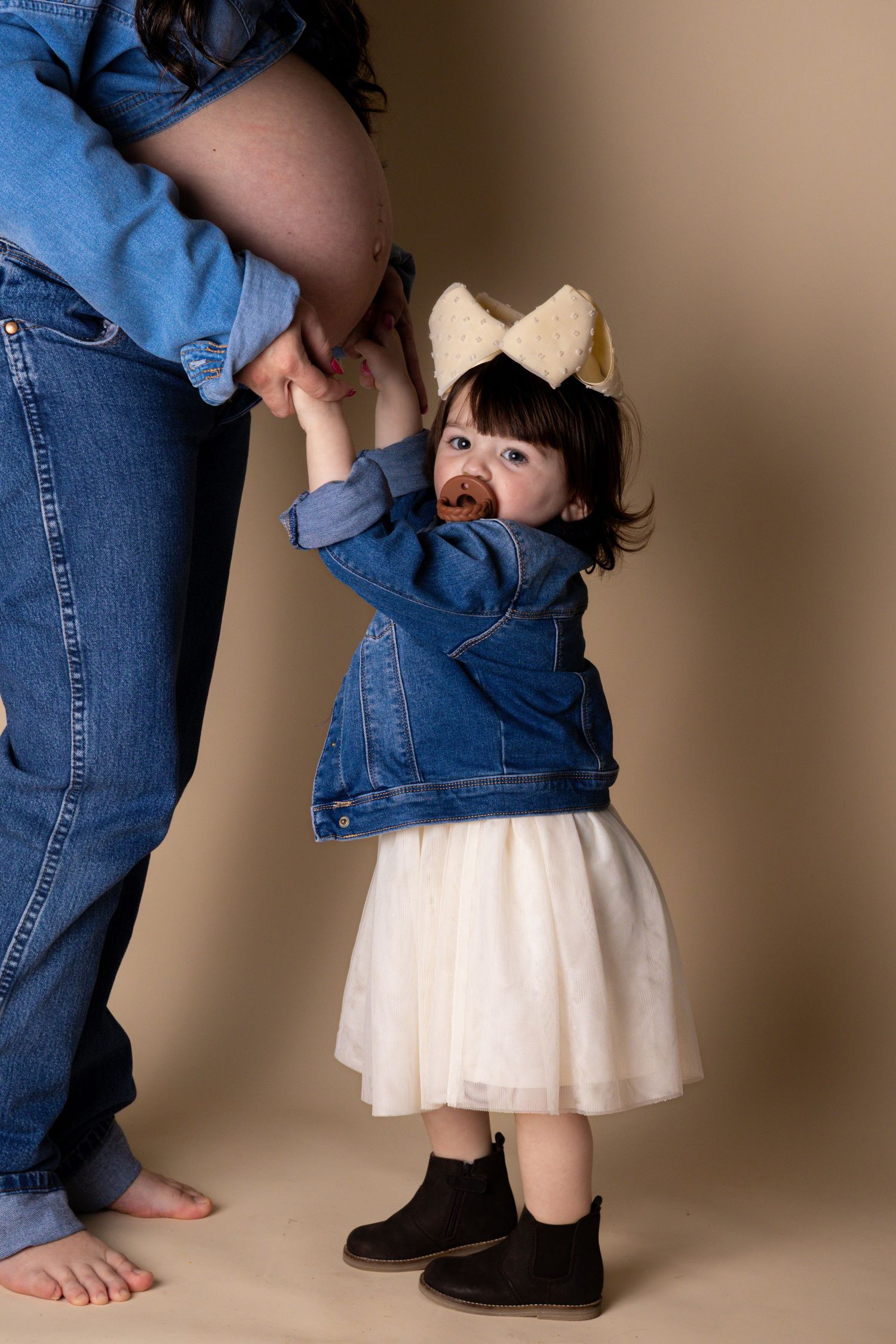 Toddler girl in denim jacket and white skirt holding hands with pregnant mother in studio.
