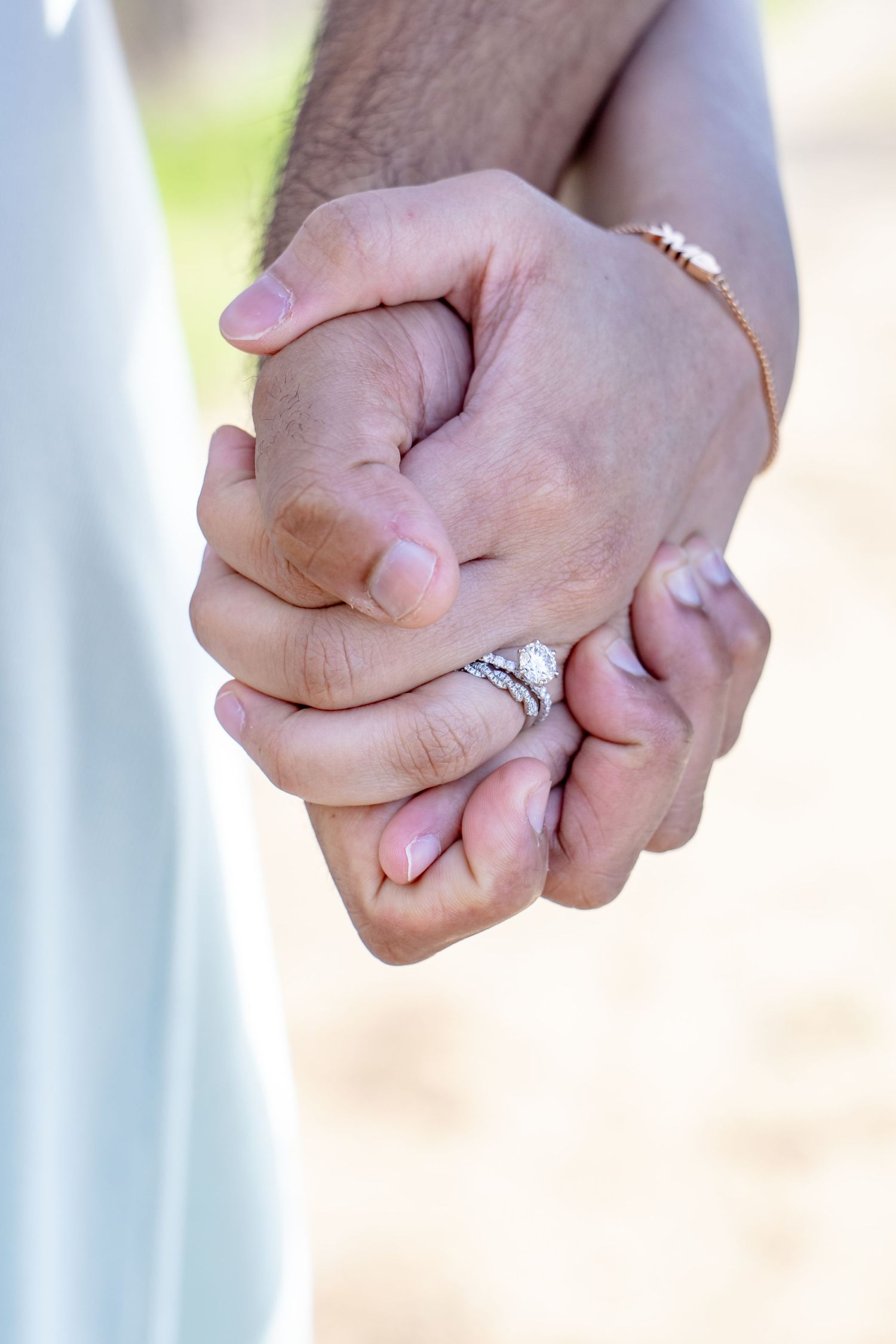Close-up of couple holding hands, woman wearing diamond engagement ring and gold bracelet outdoors.