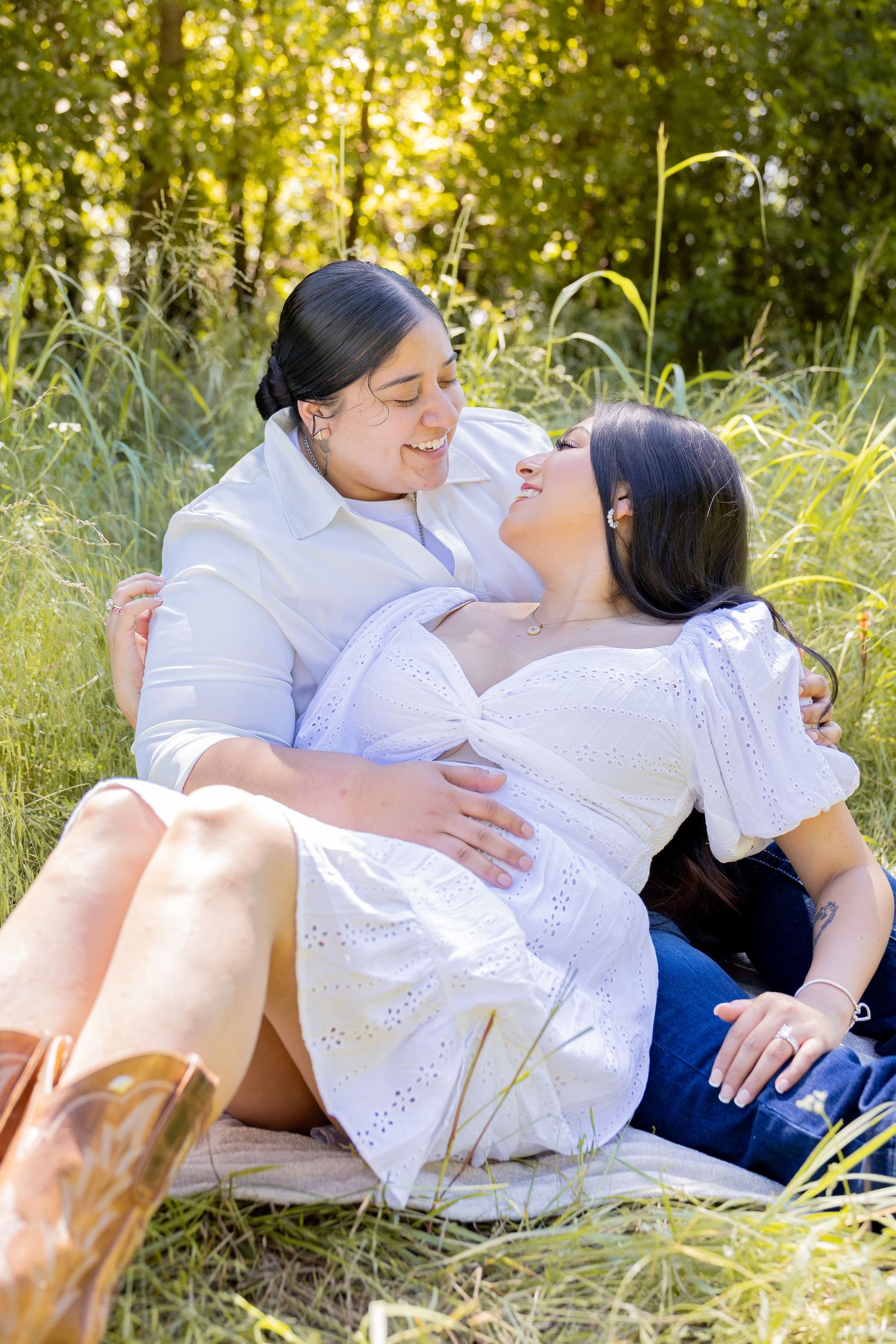 A joyful couple sits together in tall grass, smiling and embracing in a sunny outdoor meadow.