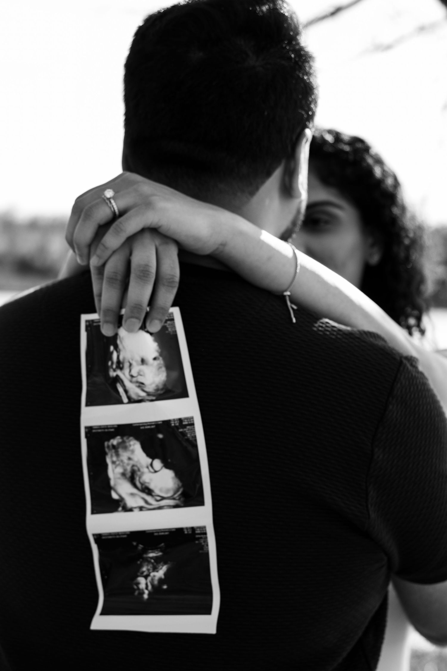 Black and white photo of pregnant woman embracing partner from behind while holding ultrasound strip images.