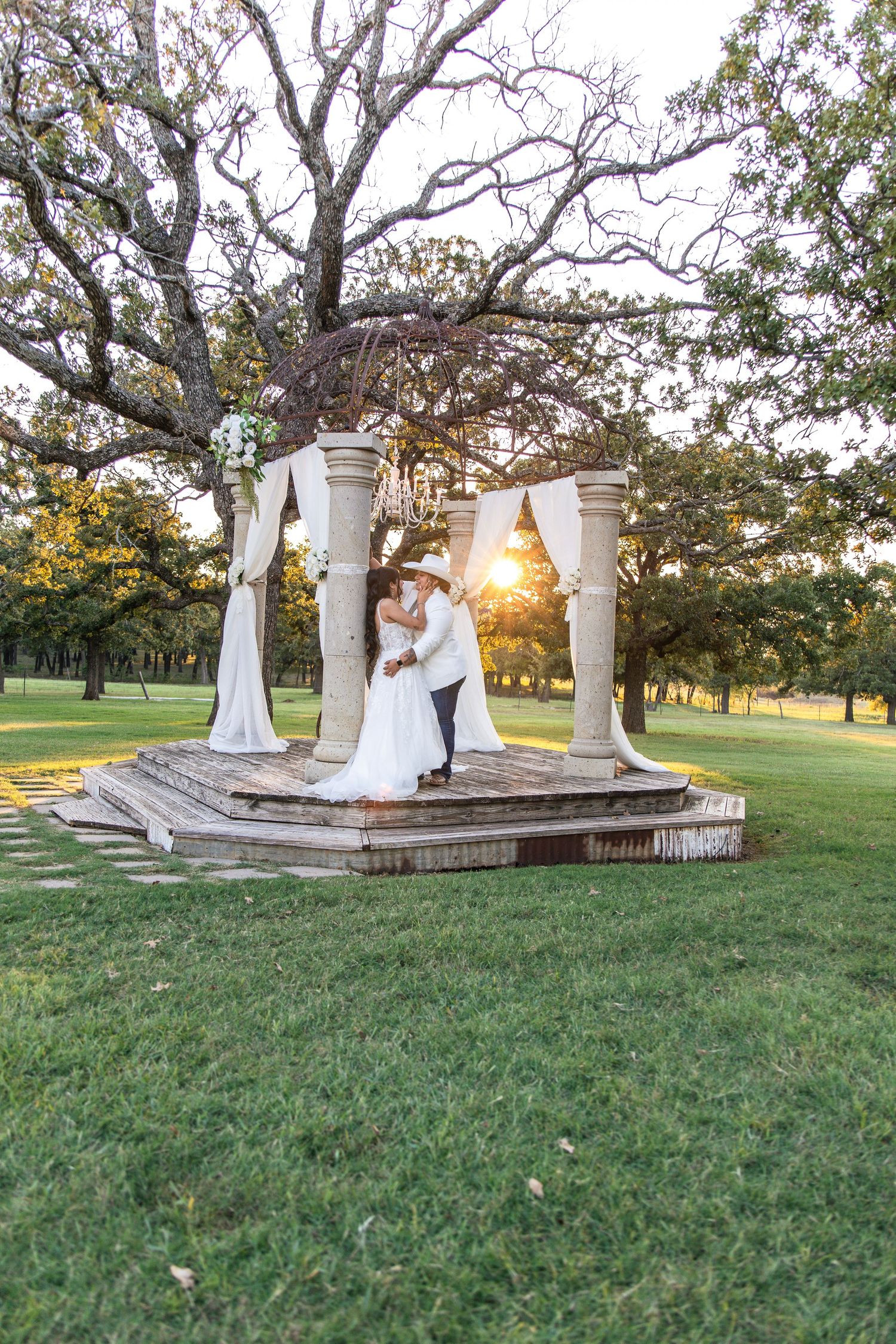 Bride and groom embrace on a stone gazebo draped in white fabric at sunset, surrounded by oak trees and green lawn.