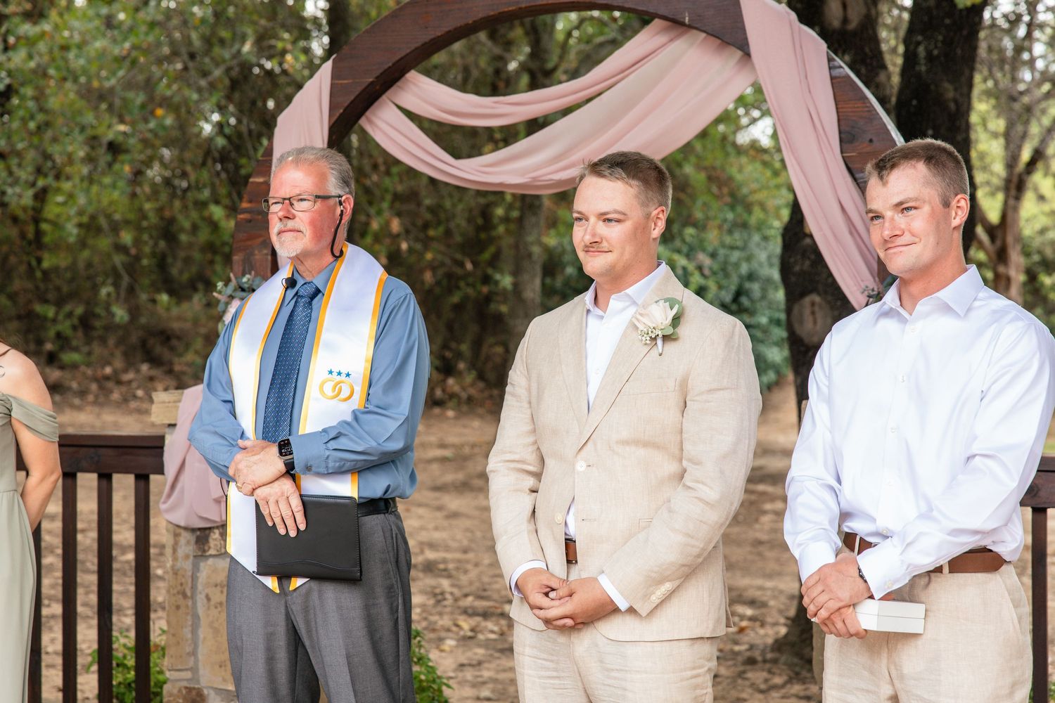 Officiant, groom in a tan suit with boutonniere, and groomsman wait at an outdoor wedding ceremony under a pink draped arch.