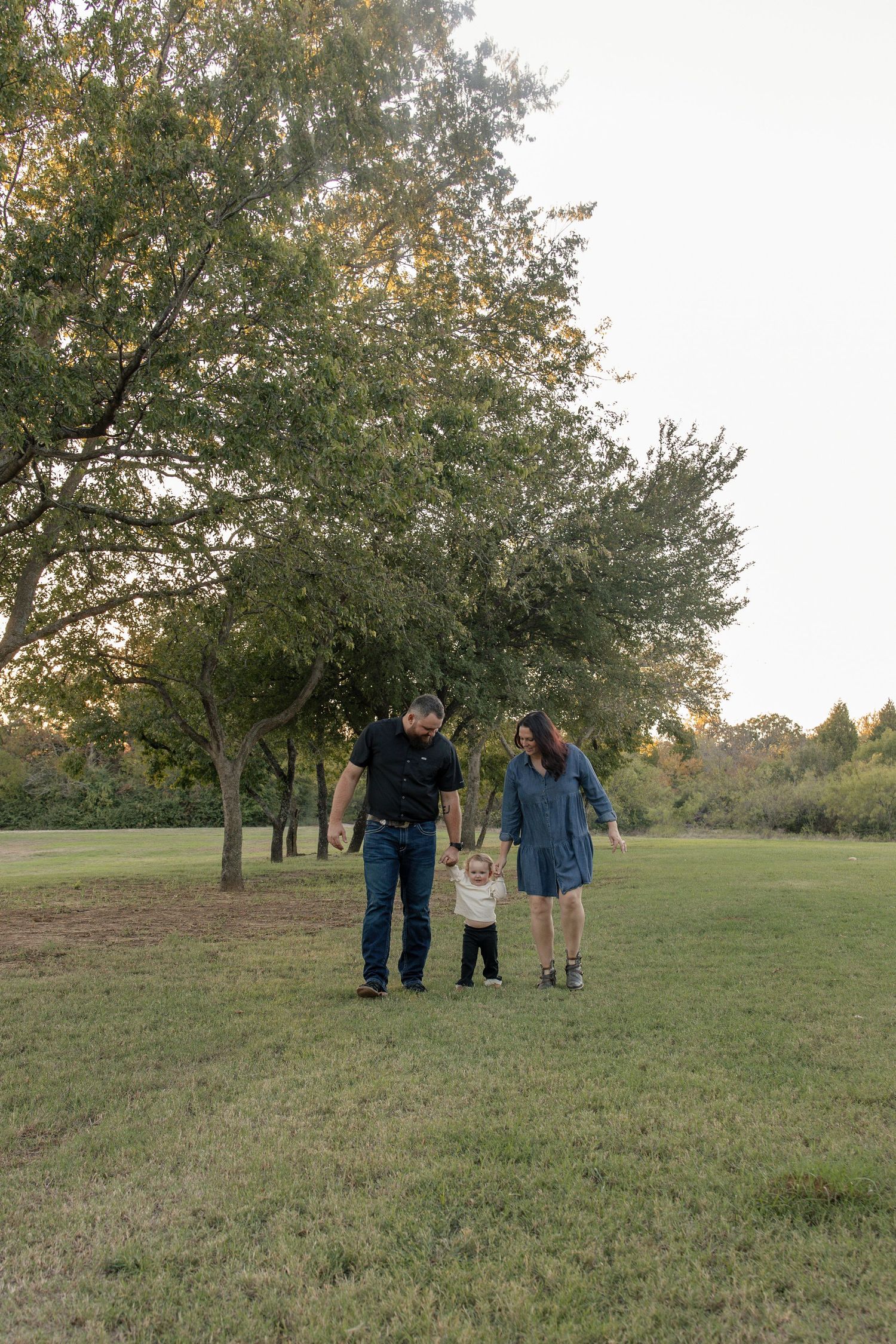 Parents walk hand in hand with their toddler across a grassy park lined with tall trees during a golden hour family photo session.