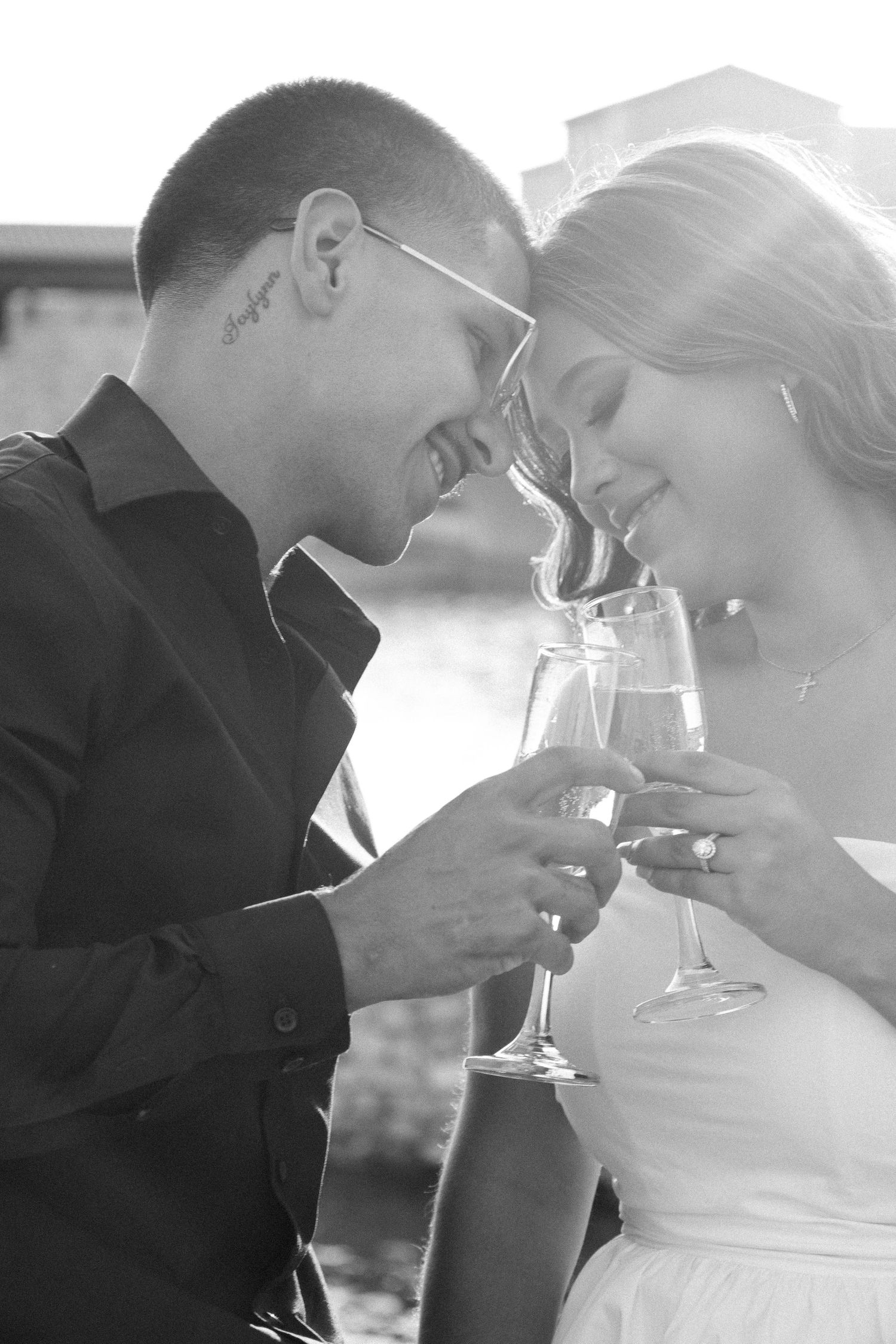 Black and white photo of newlyweds clinking champagne glasses and smiling together outdoors.