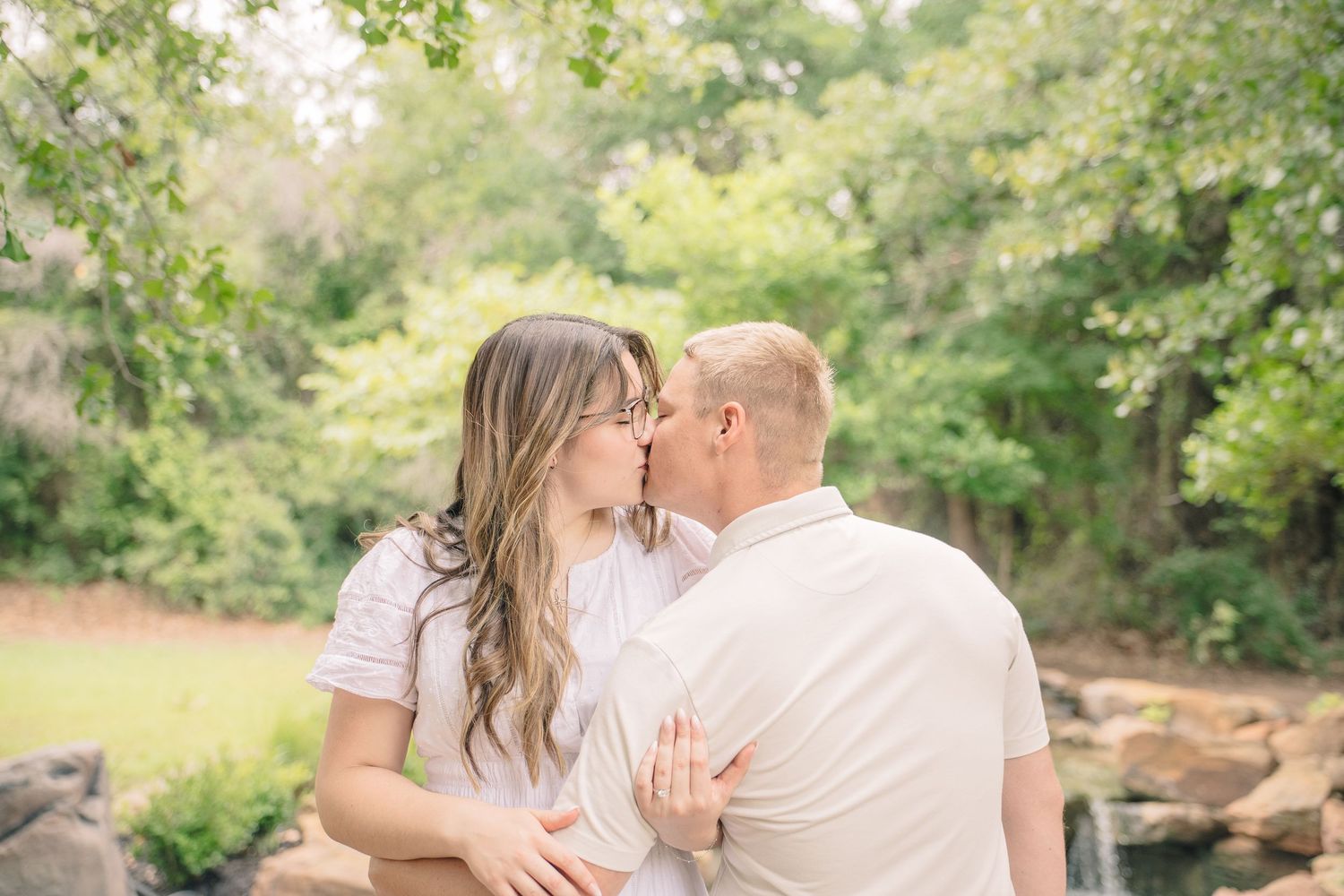 Couple sharing a romantic kiss outdoors surrounded by lush green trees and rocks in a soft natural light setting.
