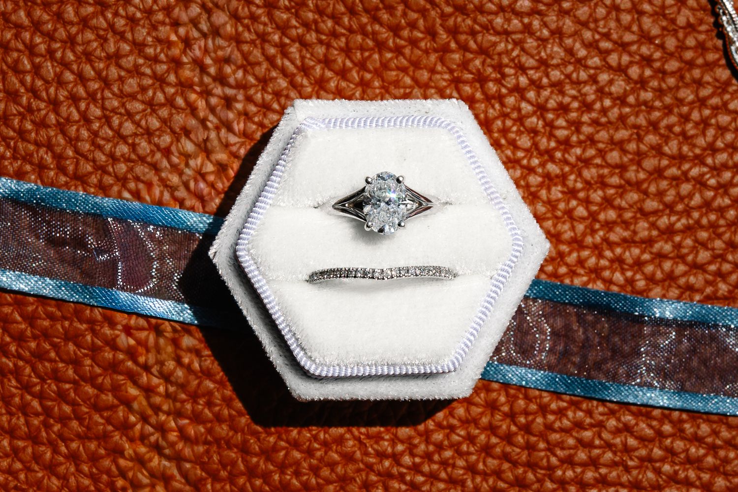 Diamond engagement ring and wedding band resting in a white hexagonal velvet ring box on a textured orange leather surface with blue ribbon.