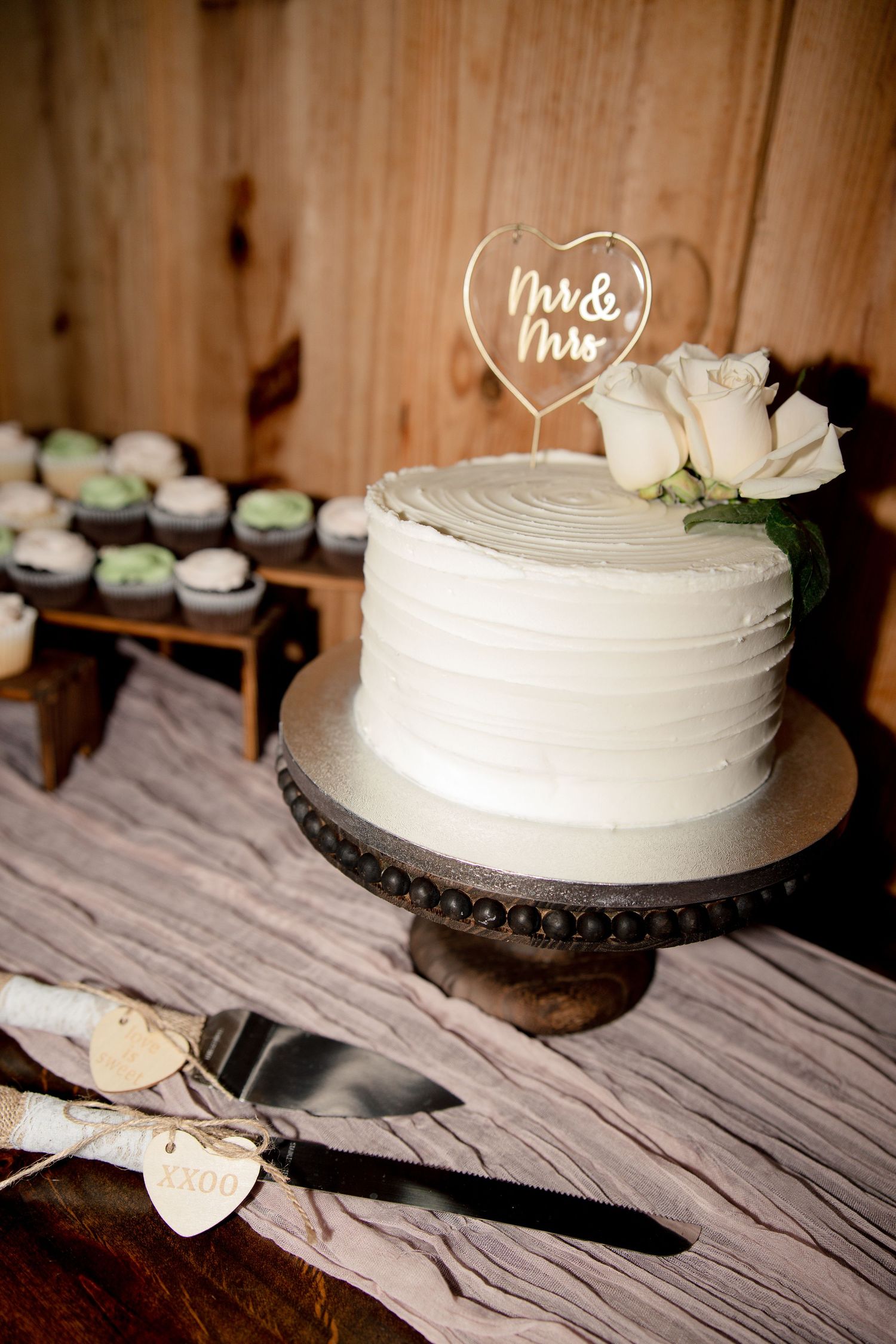 One-tier white buttercream wedding cake with white roses and a Mr & Mrs heart topper, beside cupcakes and engraved cake servers.