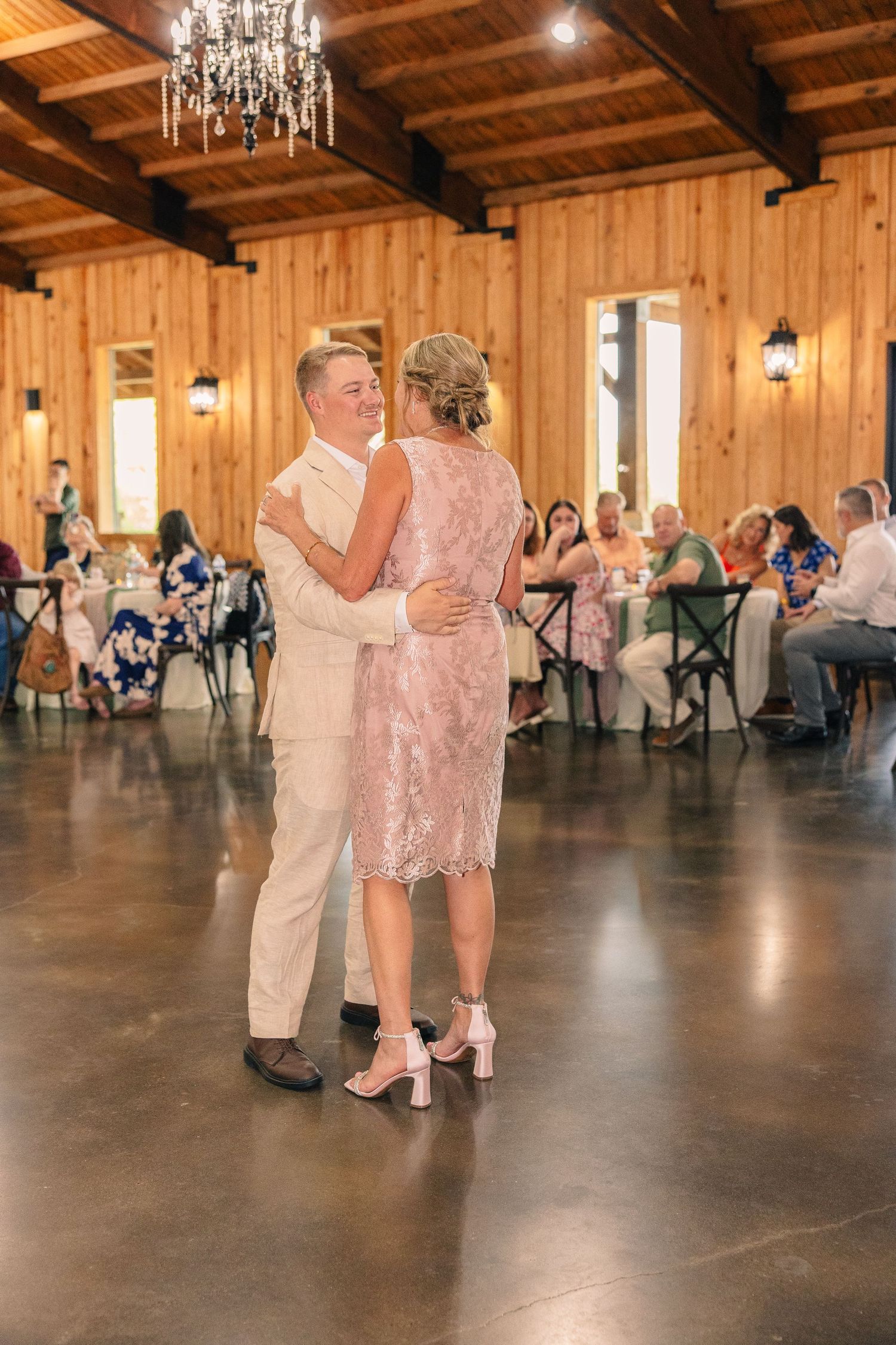 Groom dances with a woman in a pink lace dress and heels beneath a chandelier in a rustic wedding reception hall.