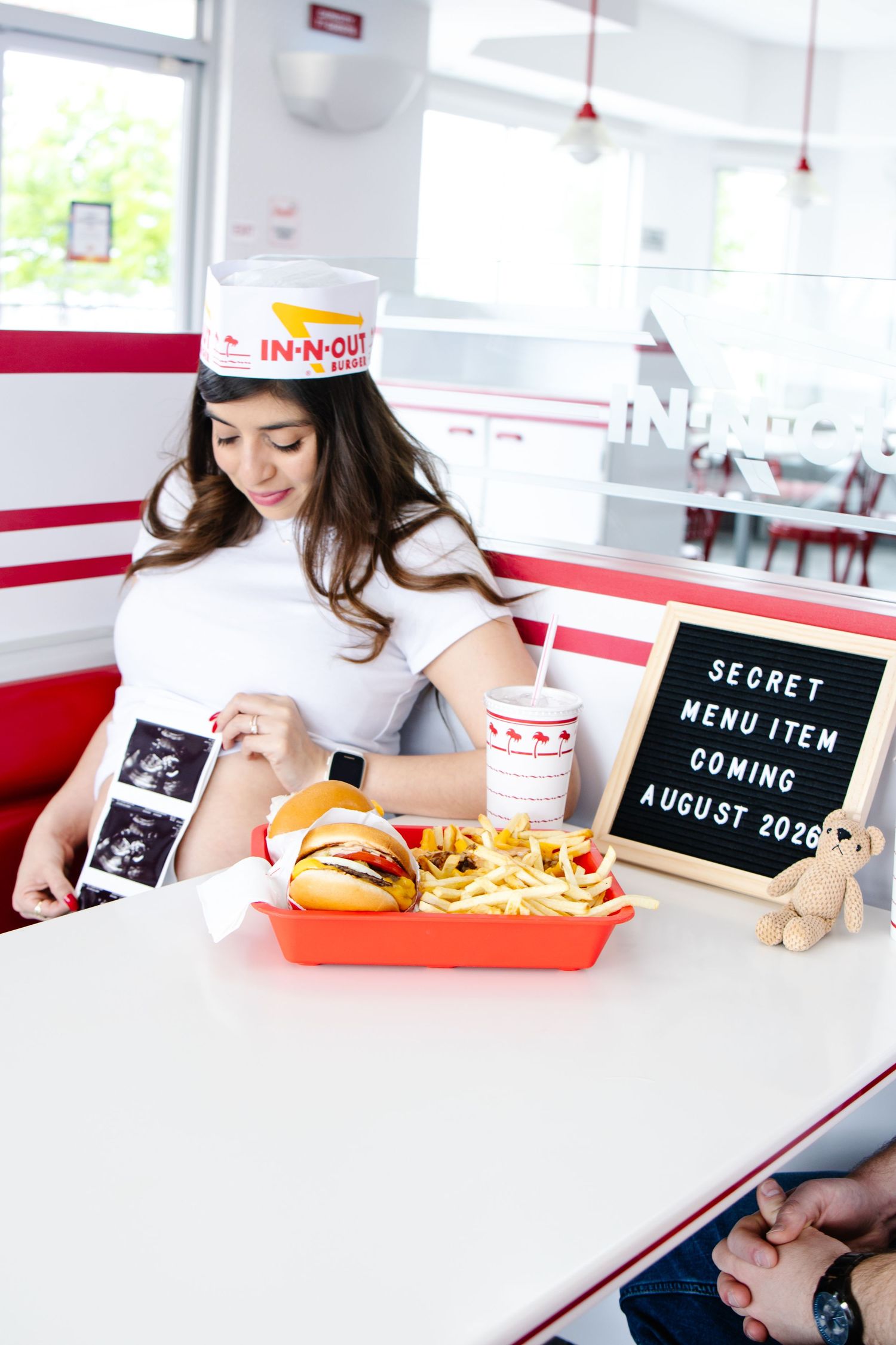Woman in an In-N-Out paper hat holds ultrasound photos beside a burger, fries and drink near a sign reading Secret Menu Item Coming August