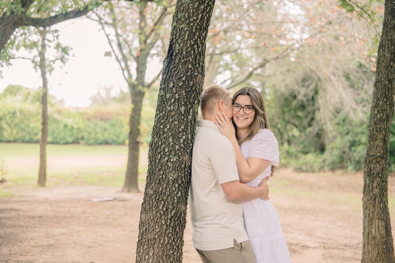 Couple embracing by tree trunks in a park, woman in white dress smiling, man in beige shirt, soft natural light.