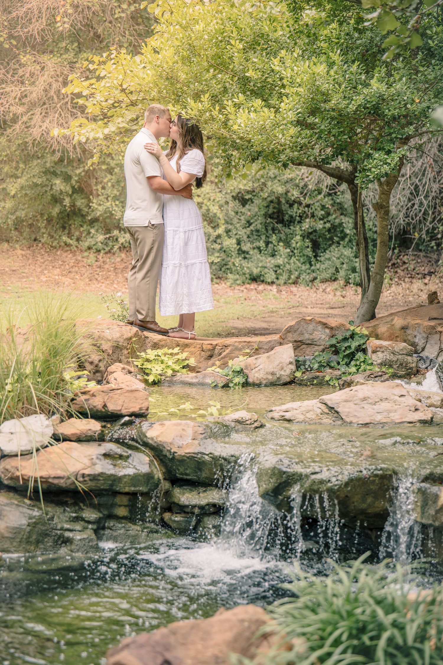 Couple sharing a romantic kiss on rocks beside a serene garden waterfall surrounded by lush green trees.
