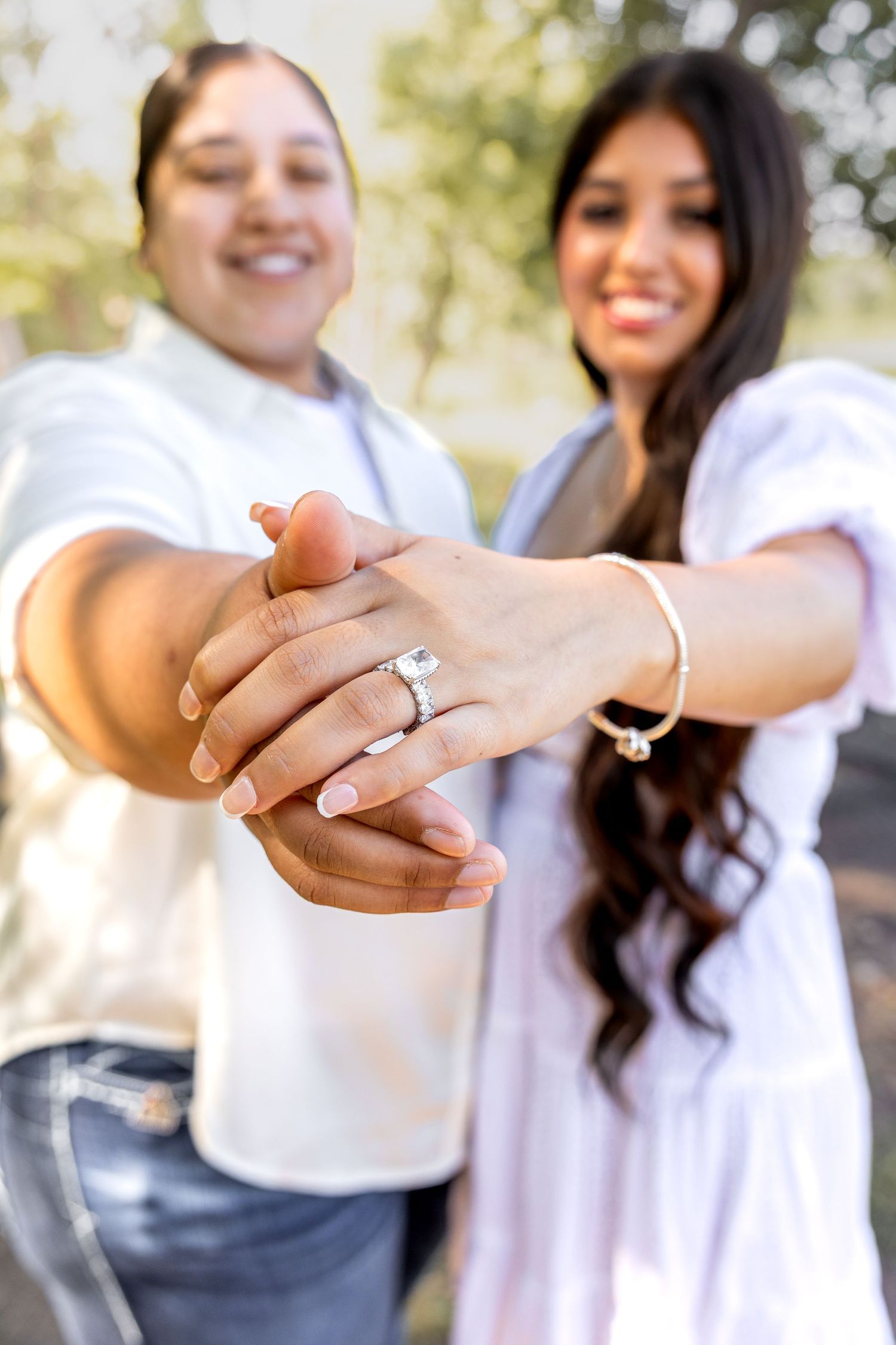 Two women smiling and showing off an engagement ring outdoors in a park setting.