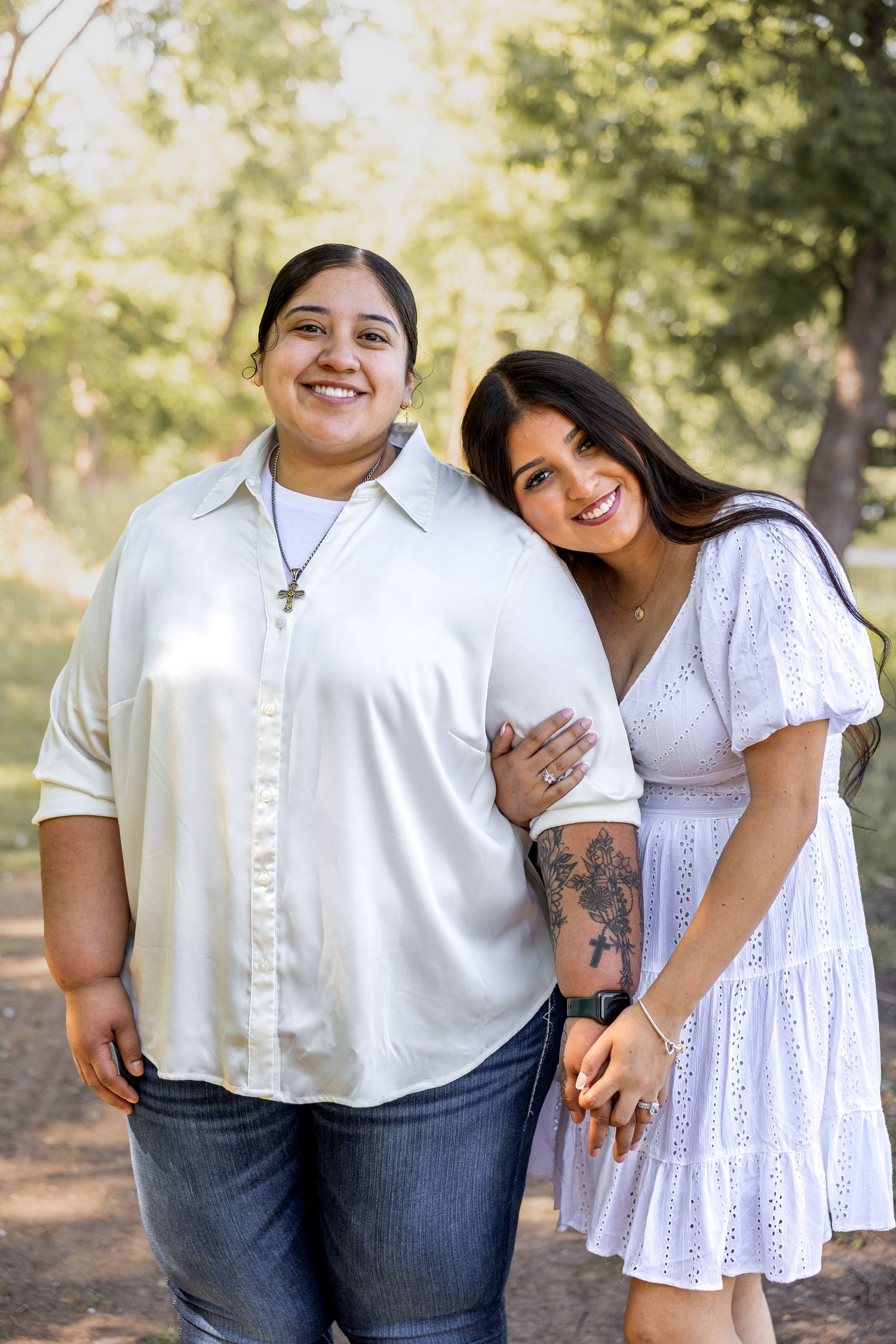 Two smiling women posing together outdoors, one in a white button-up shirt and jeans, the other in a white dress.