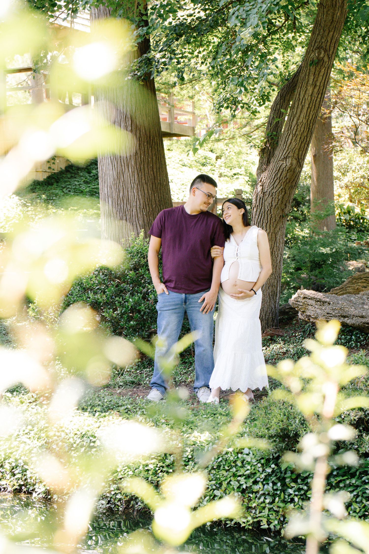 Couple sharing tender moment under trees, pregnant woman in white dress with partner in maroon shirt in lush garden.