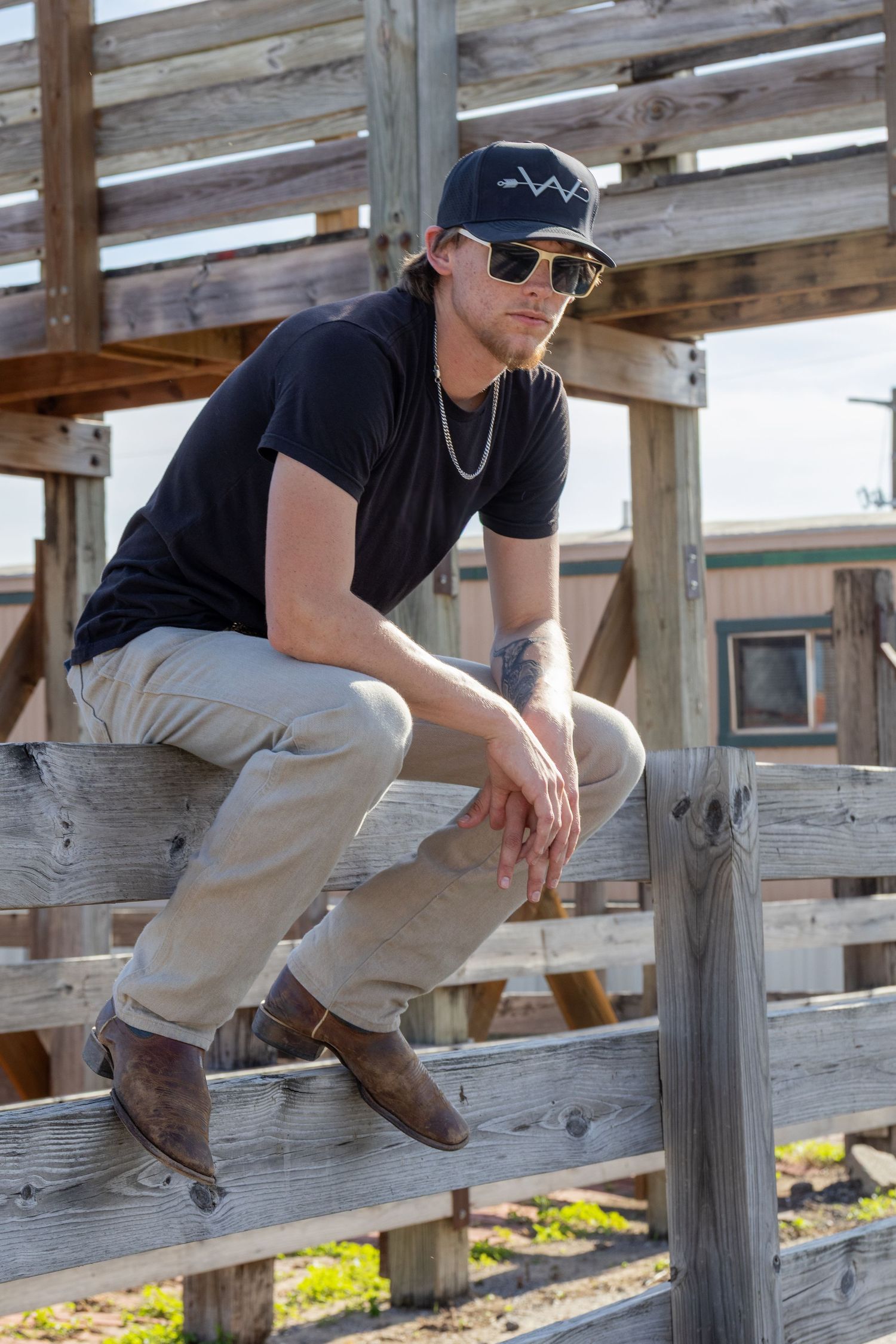Young man in black cap, sunglasses, and brown boots posing on rustic wooden fence.