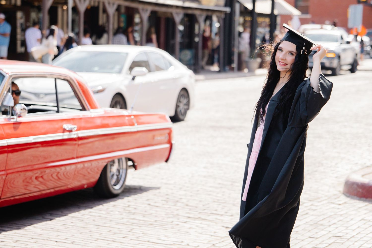 Graduate in black cap and gown posing on brick street beside a classic red vintage car.