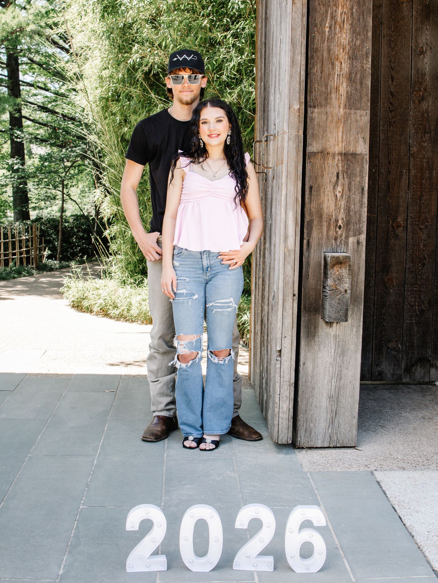 Couple standing by a wooden door next to a 2026 sign in a garden setting.
