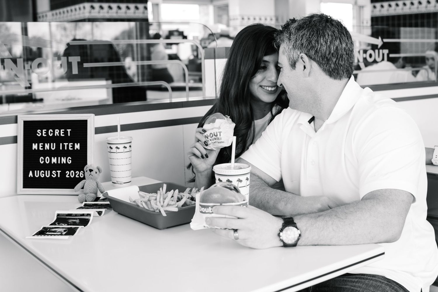 Black and white photo of couple at In-N-Out with pregnancy announcement letter board on the table.