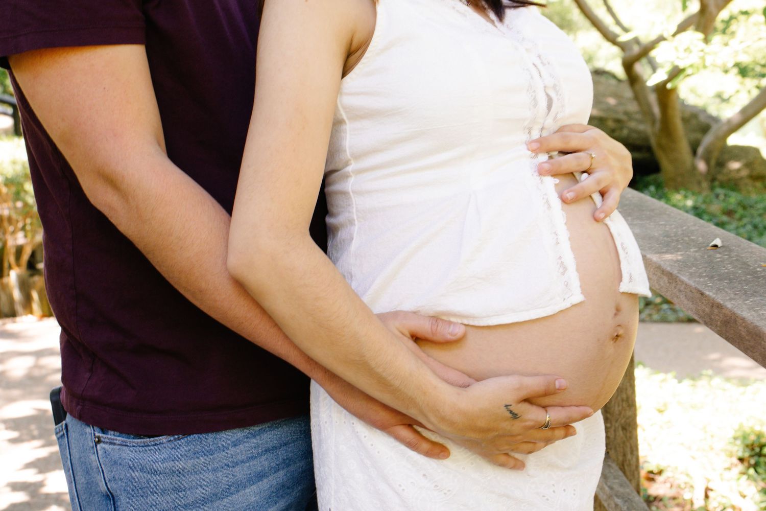 Close-up of couple embracing pregnant belly outdoors during sunny maternity photo session in park.
