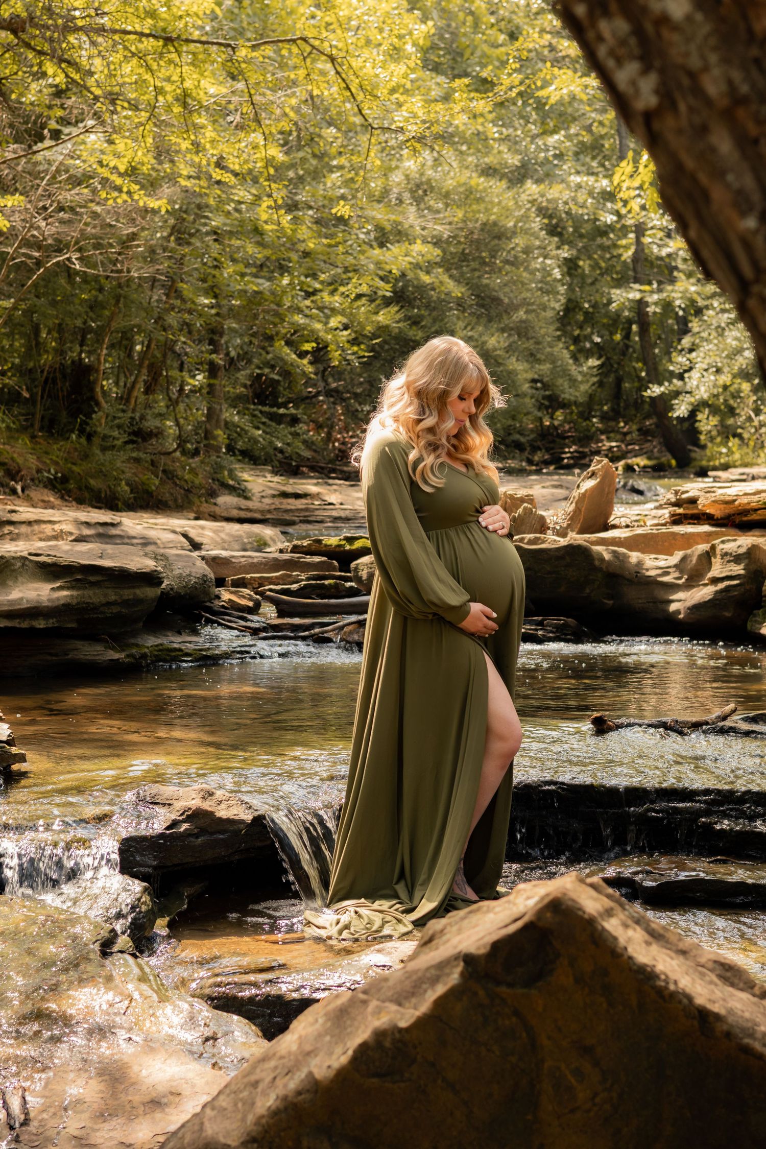 Pregnant woman in flowing olive green gown standing on rocks beside a peaceful forest stream.