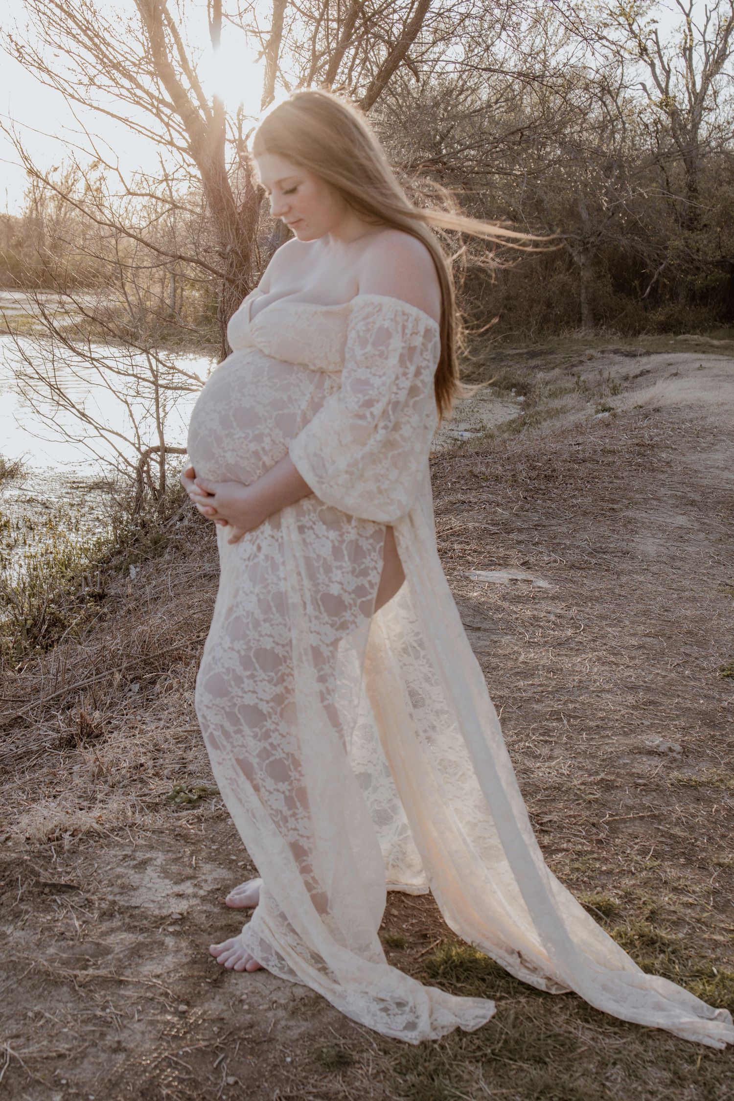 Pregnant woman in white lace gown standing by a river at golden hour sunset.