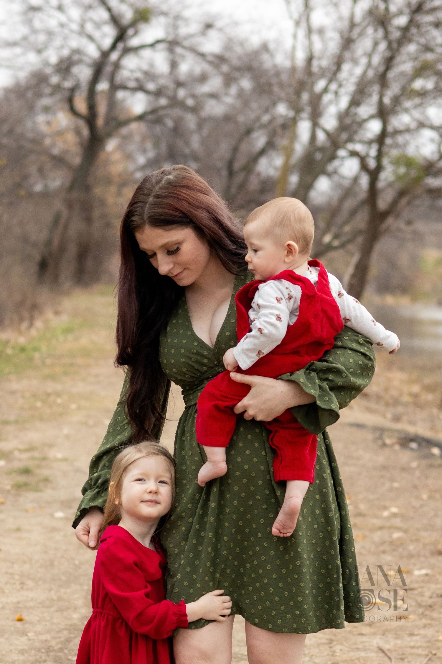 A young mother in a green dress holds her baby while her toddler daughter in red clings to her.