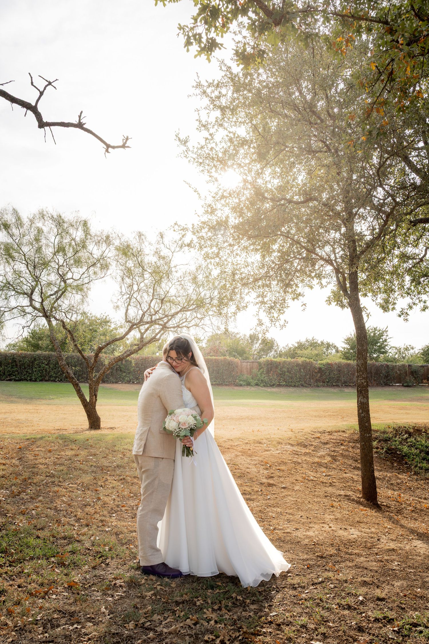 Bride and groom embracing in golden sunlight among trees, bride holding white bouquet in flowing white gown.