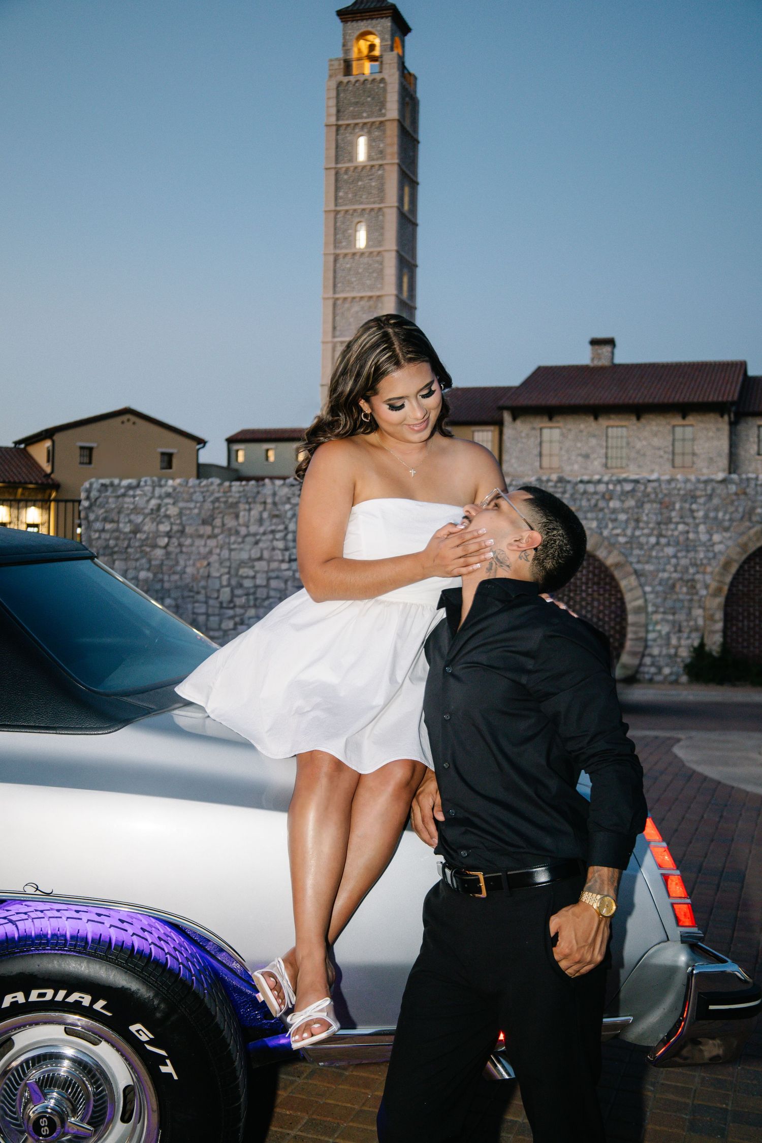 Couple posing with classic white car at dusk, bride in white dress sitting on hood while groom looks up adoringly.