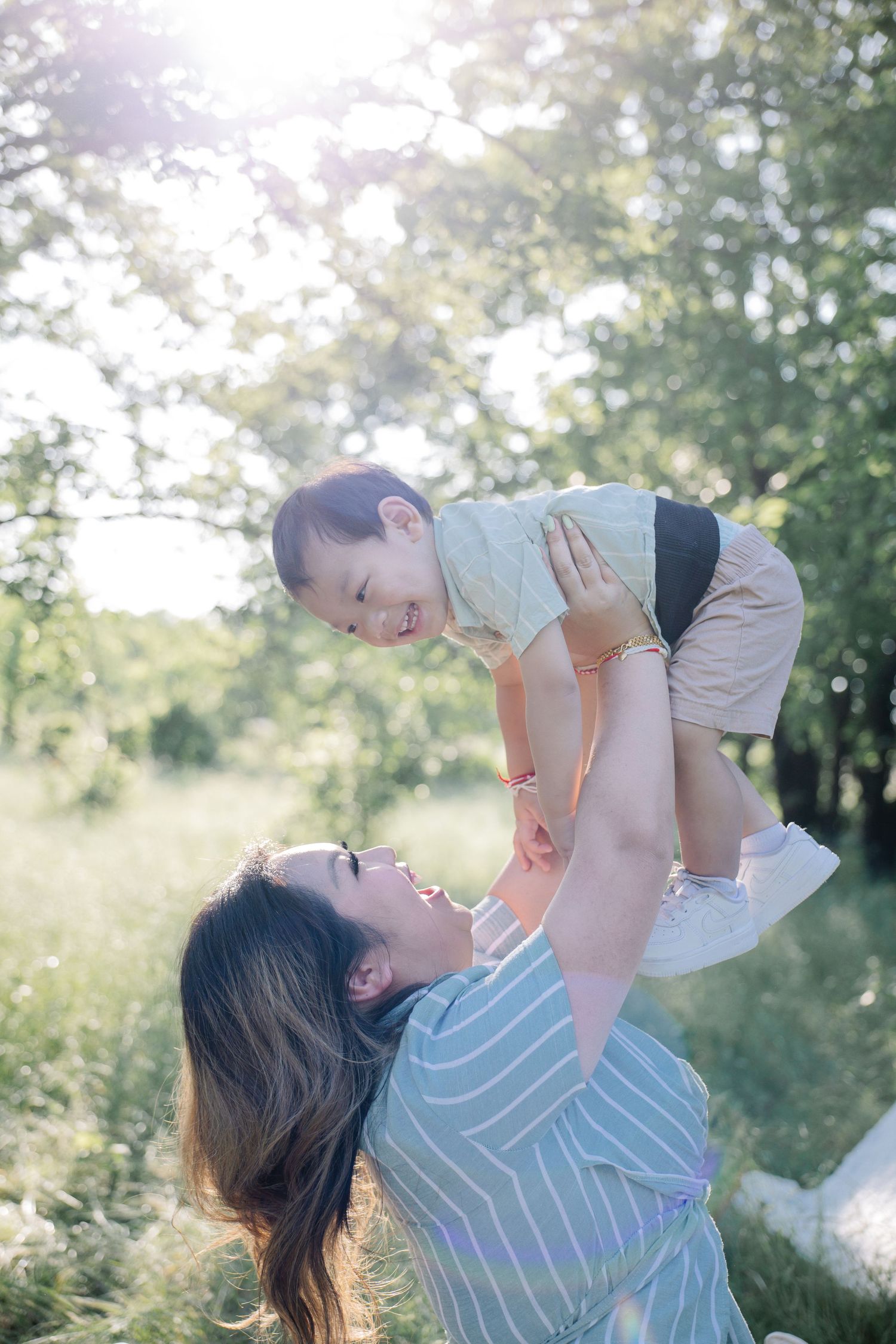 A joyful mother lifts her smiling toddler high in a sunlit green park, sharing a playful and loving outdoor moment.
