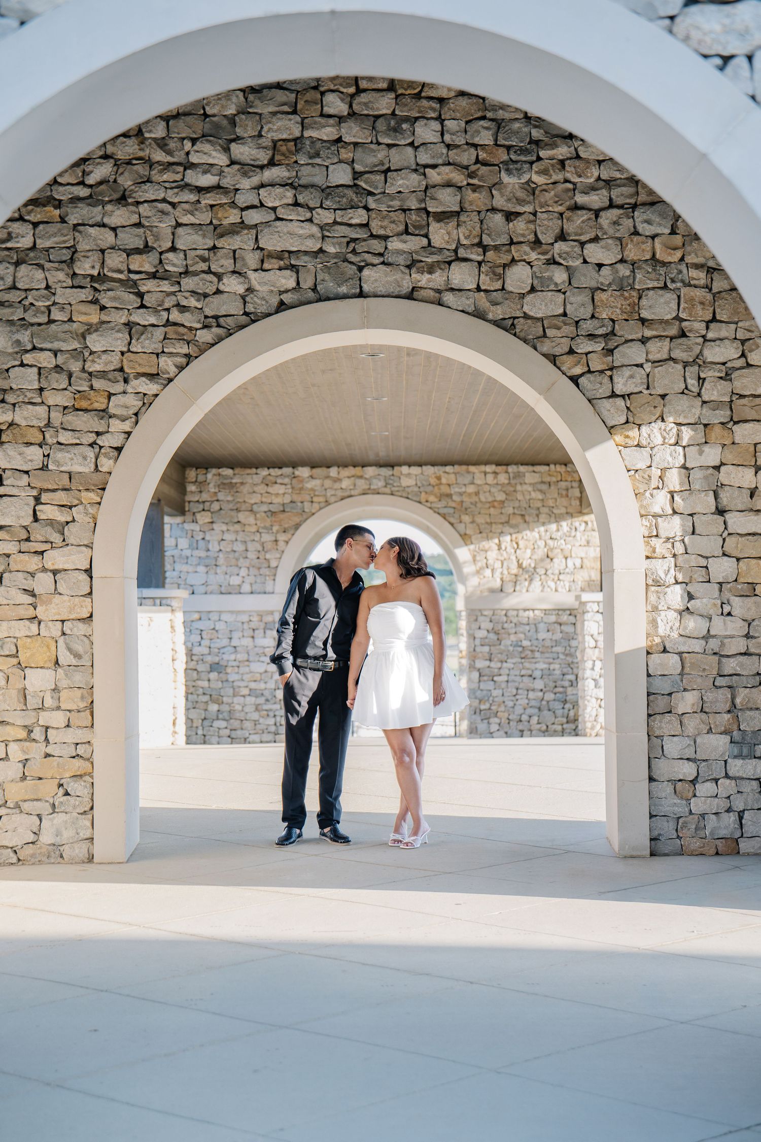 Couple sharing a romantic kiss under white stone arches at an outdoor wedding venue.