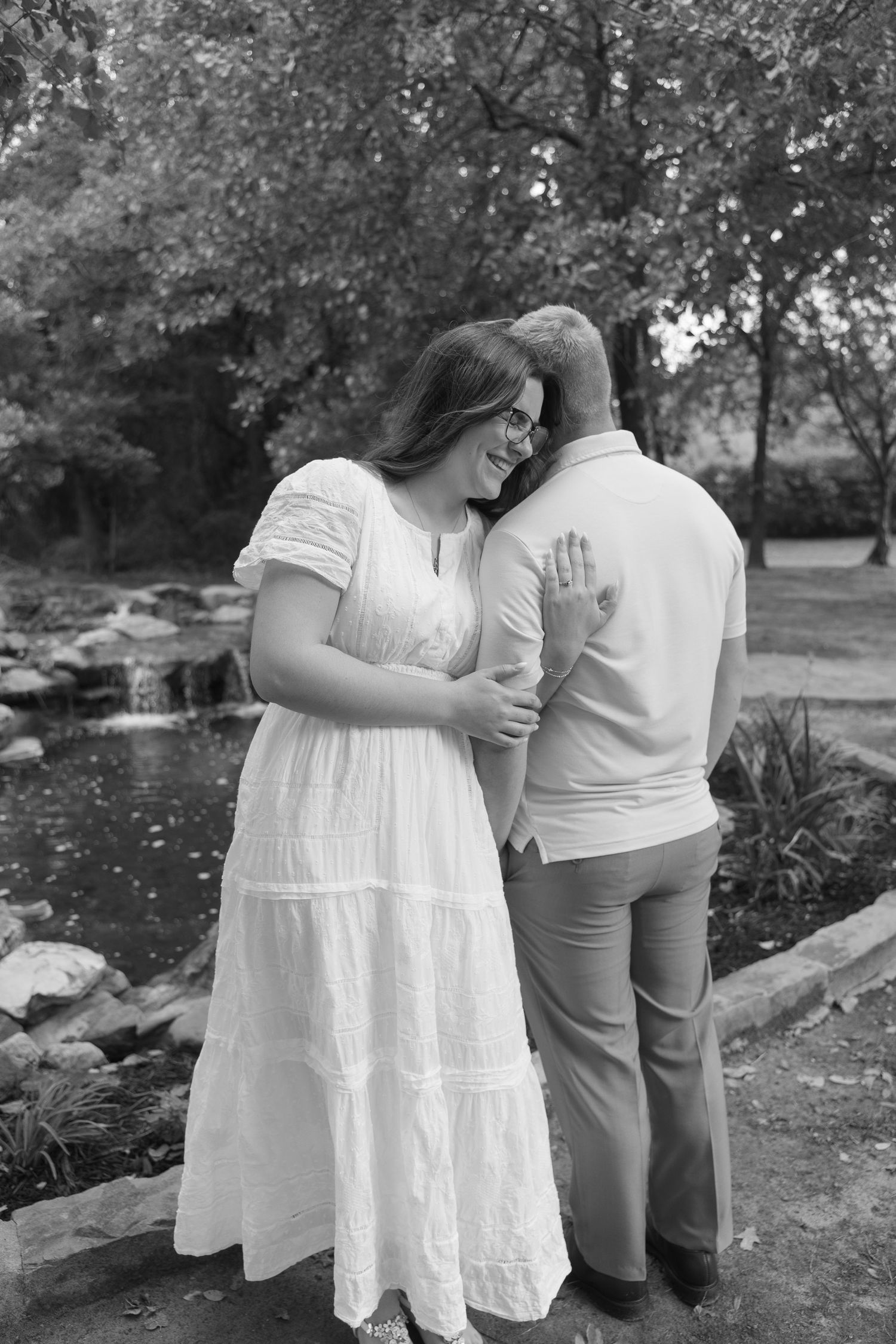 Black and white photo of a couple embracing near a pond in a garden, woman wearing a white dress showing her engagement ring.