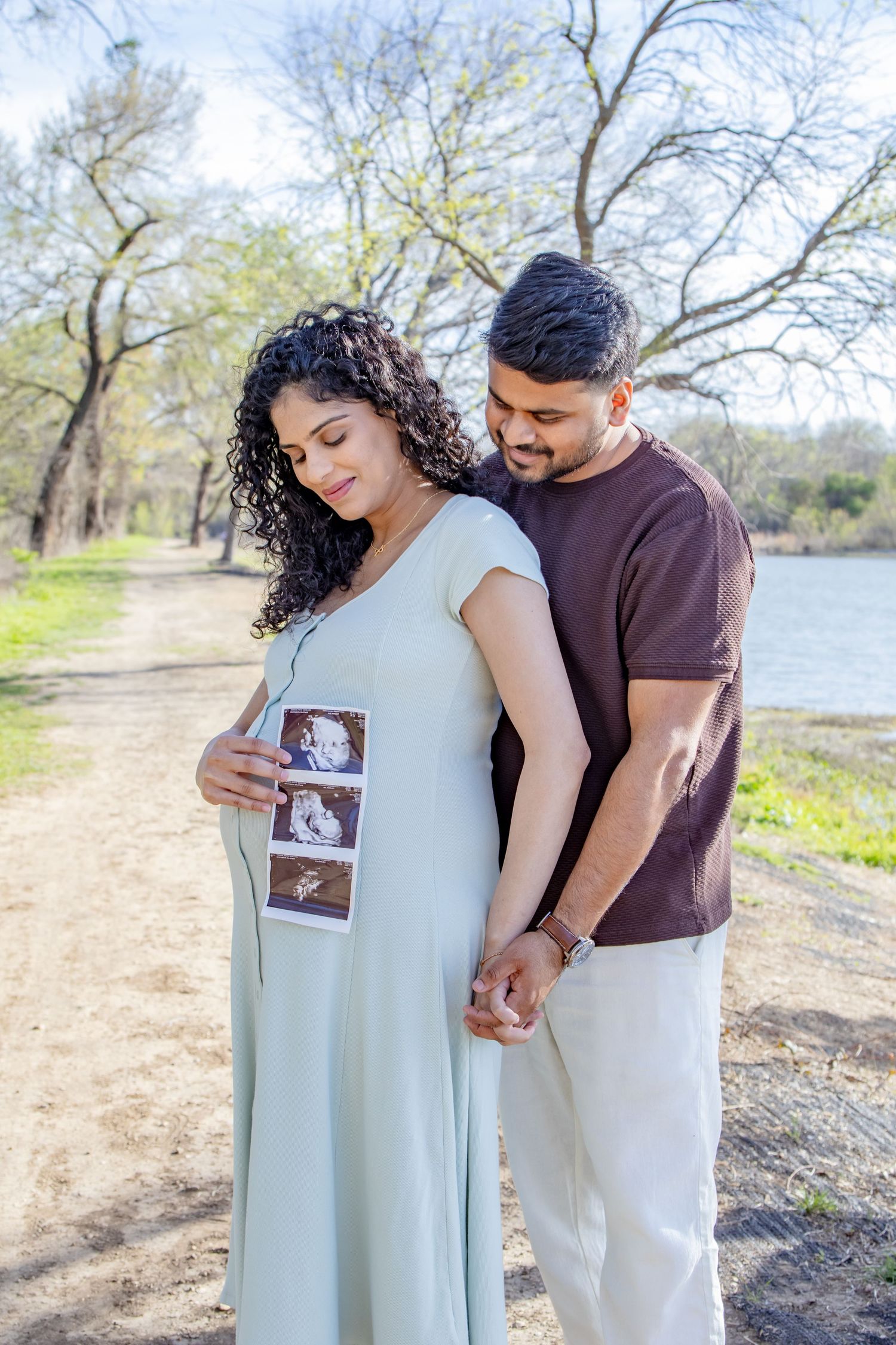 Pregnant couple holding ultrasound photos outdoors on a sunny path, man embracing woman from behind while holding hands.
