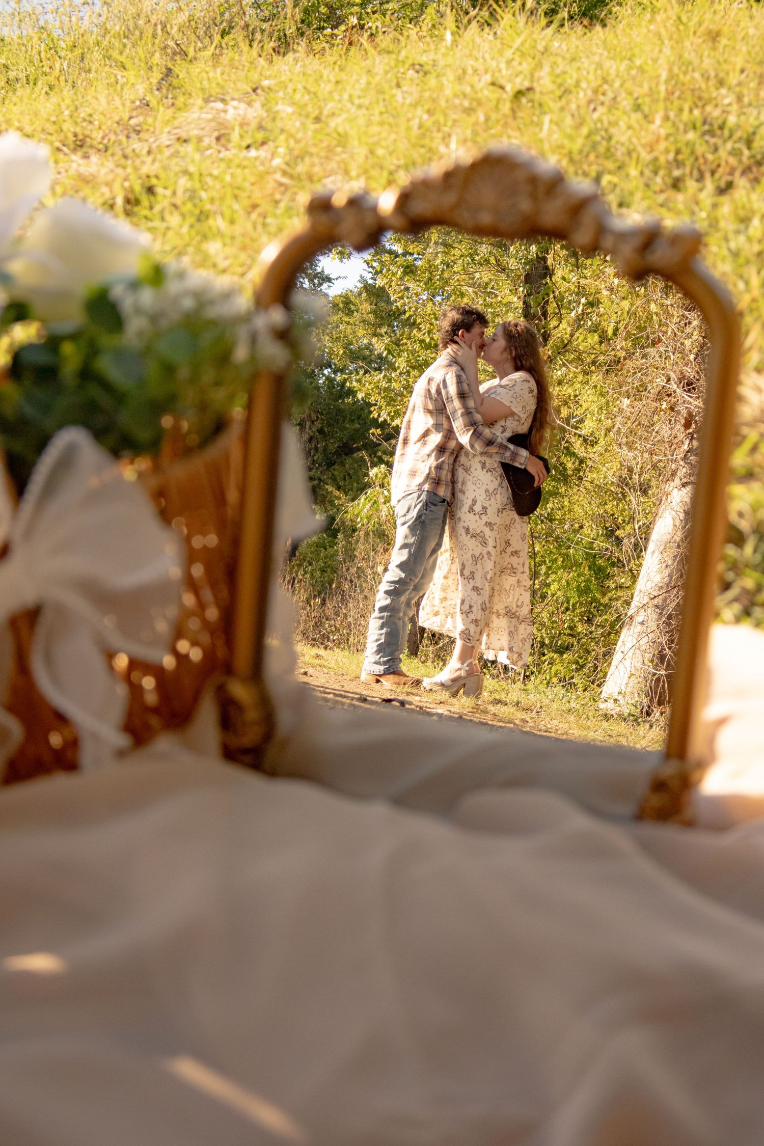 Romantic couple embracing outdoors reflected in a gold vintage mirror with a white floral arrangement in the foreground.