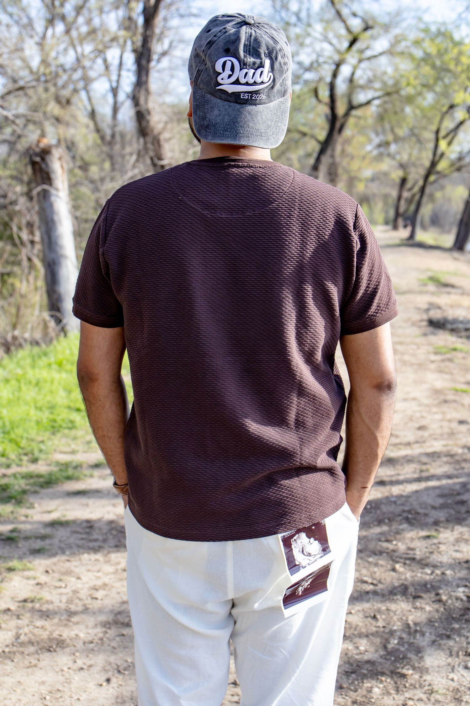 Expectant father walking away on a nature path wearing a gray Dad cap with ultrasound in pocket.