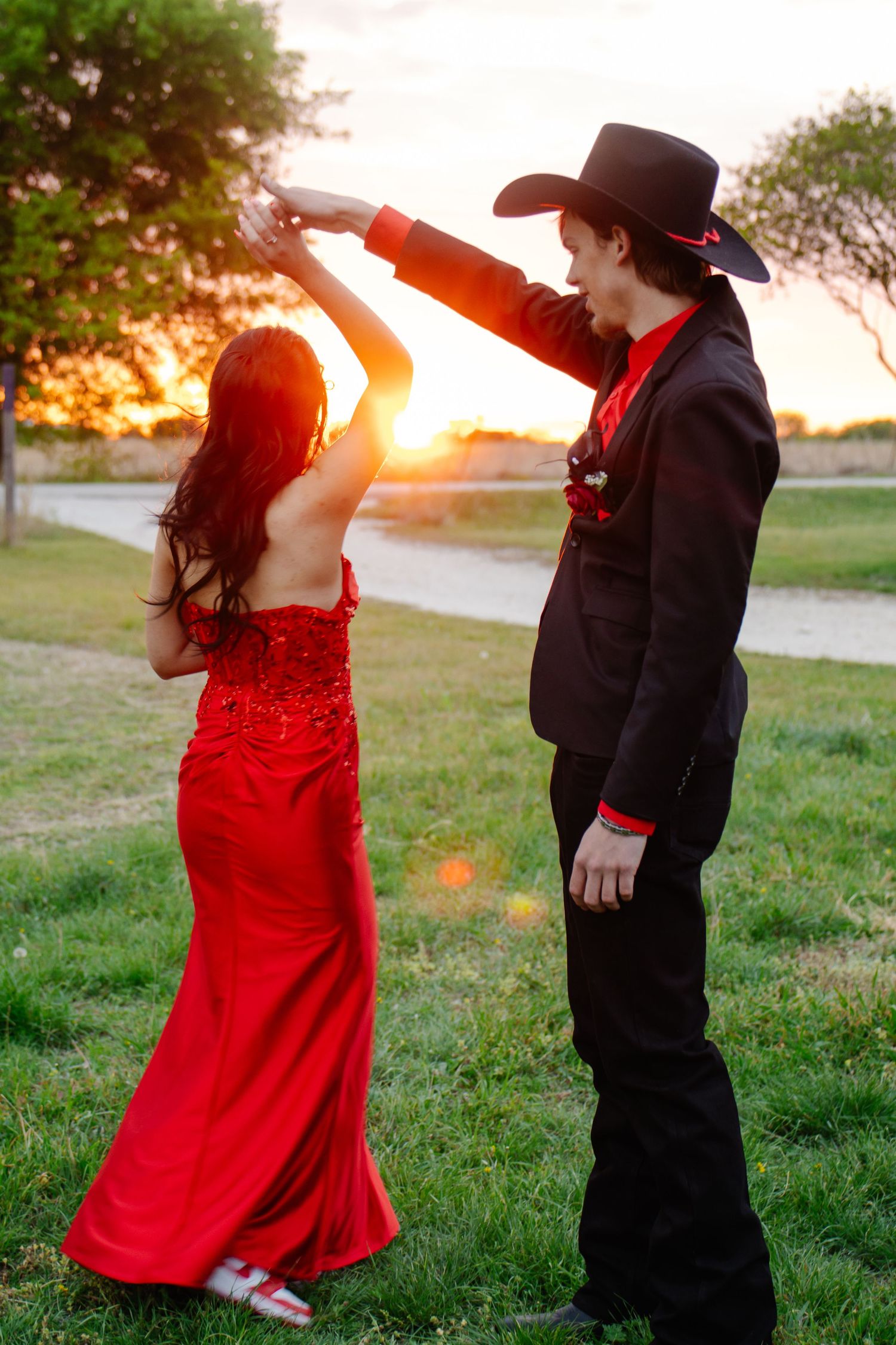 Prom couple dancing at sunset, boy in black cowboy hat spinning girl in red sequin gown outdoors.