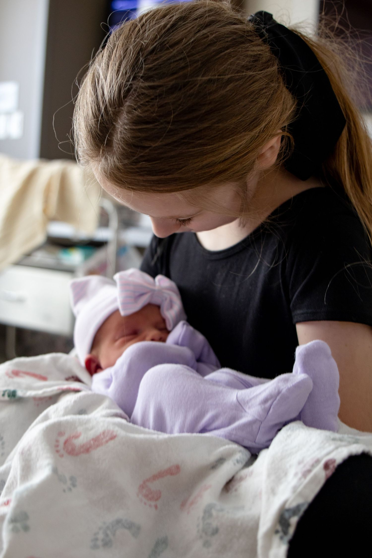 Young girl with ponytail tenderly holding and gazing at her newborn sibling in purple outfit.