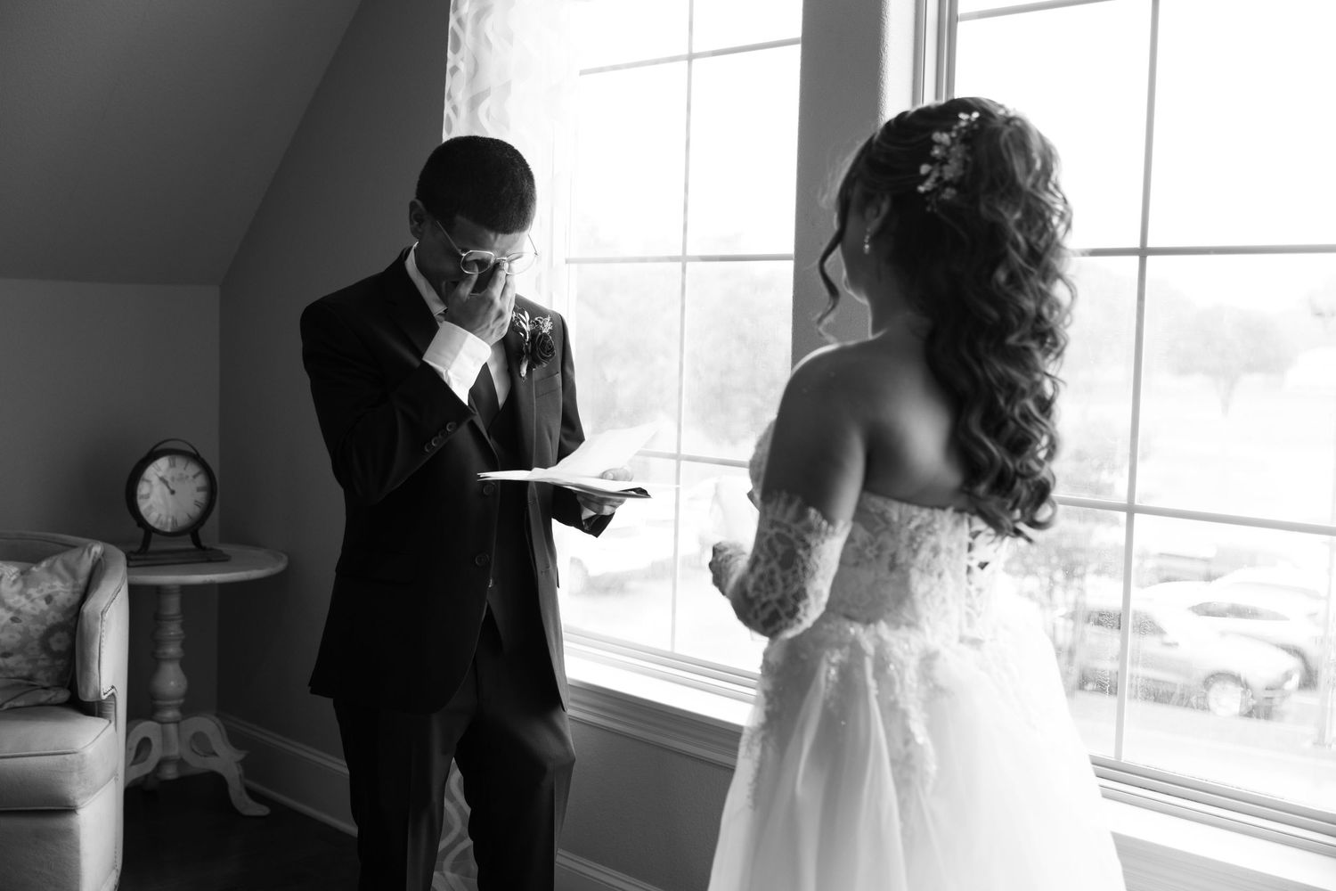Groom reads vows while bride listens by window in emotional black and white wedding moment.
