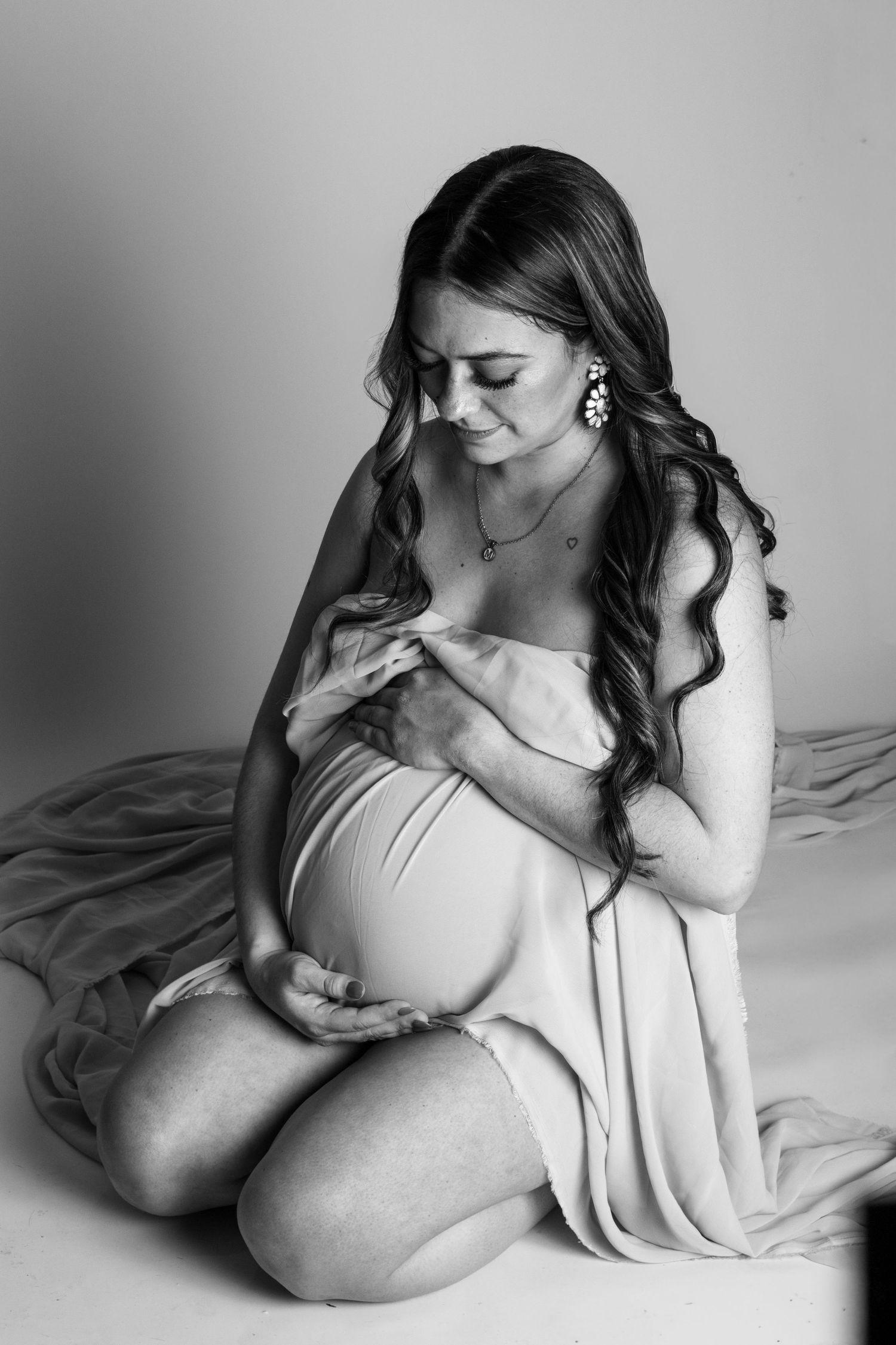 Black and white maternity photo of a pregnant woman kneeling, cradling her baby bump while smiling softly.